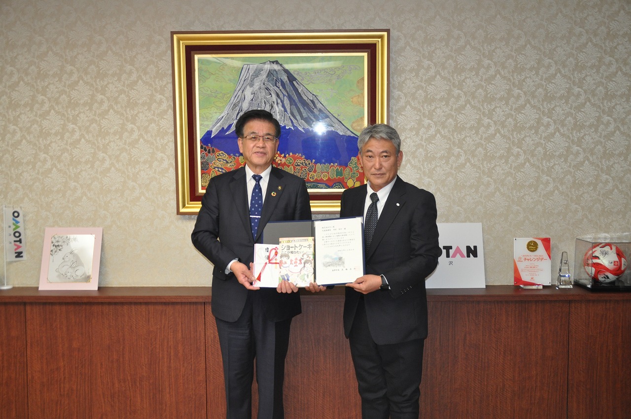 富士山の絵を背景に高橋市長と菊池工場長が寄贈された本と寄贈を示す証書を持ち、笑顔で写っている記念写真