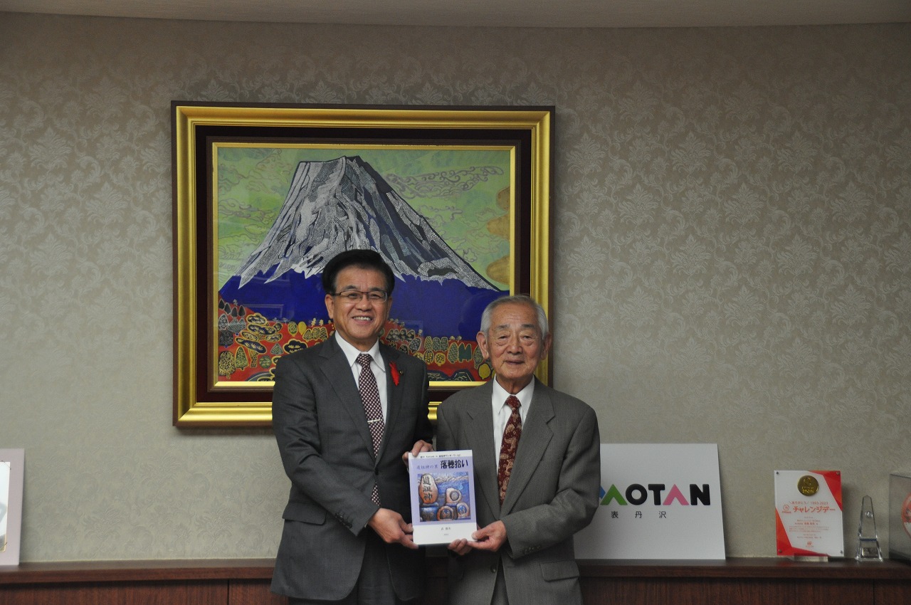 富士山の絵を背景に、スーツ姿の武勝美さんと高橋市長が寄贈された本を持って笑顔で写っている記念写真