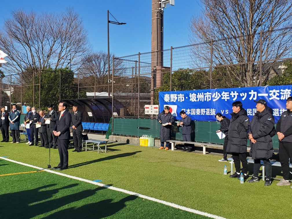 晴天の下、「秦野市・坡州市少年サッカー交流試合」と書かれた横断幕が掲げられた会場で、スーツ姿の来賓やコートを着た関係者たちが整列し、中央で高橋市長がマイクの前でスピーチをしている開会式の様子の写真