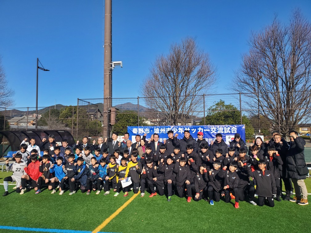 「秦野市・坡州市少年サッカー交流試合」と書かれた横断幕の前で、試合に参加する両市の少年サッカーの選手たちと、スーツ姿の来賓やコート姿の関係者たちが笑顔で整列し、全員で拳を掲げながら写っている集合写真