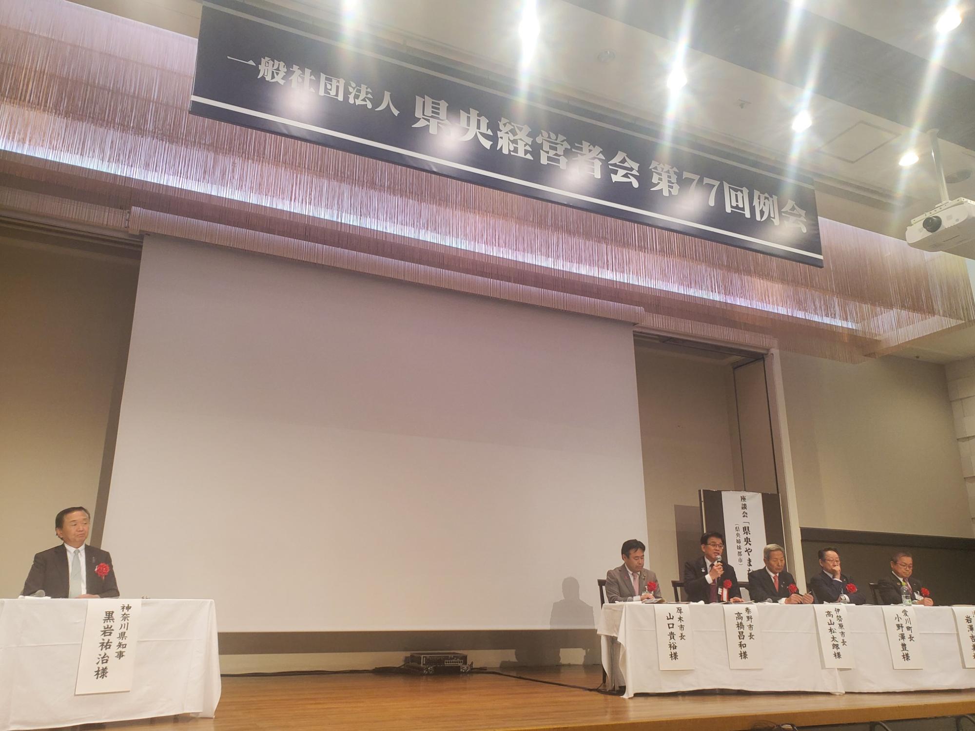 「一般社団法人 県央経営者会 第77回例会」と書かれた垂れ幕の下、舞台上には神奈川県知事をはじめとする複数の自治体市長や関係者が横一列に座り、マイクを使って発言している様子
