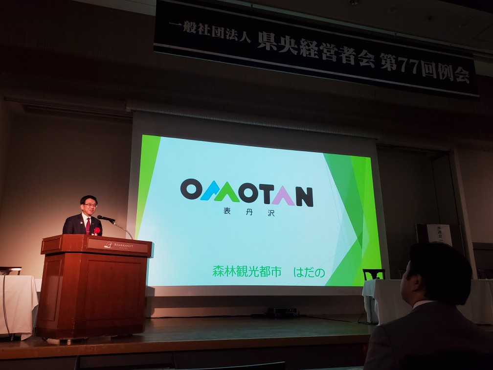 『OMOTAN（表丹沢） 森林観光都市 はだの』と書かれたスライドを背に、壇上に立つ高橋市長がマイクの前で話をしている様子の写真