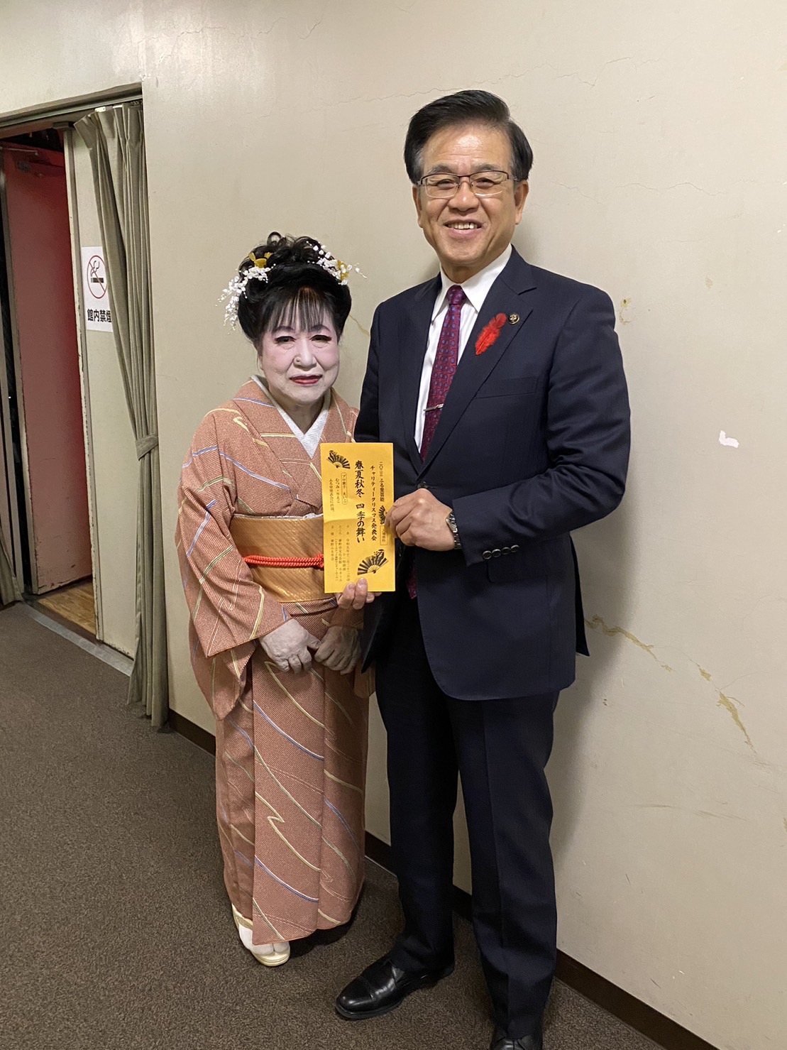 和装姿の女性とスーツ姿の市長が壁際で並び、舞台出演の案内状を手に笑顔でカメラに向かって立っている記念写真