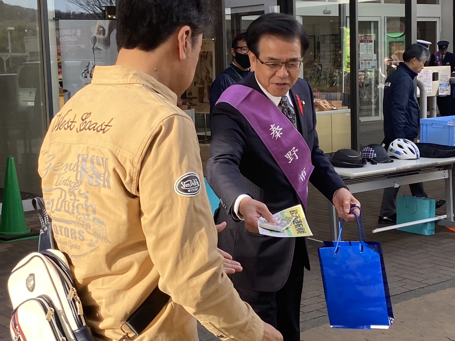 「秦野市」と書かれた紫色のタスキを肩にかけた高橋市長が、街頭に立ち、市民の男性にチラシなどを手渡ししている交通事故防止・防犯キャンペーンを行っている様子の写真
