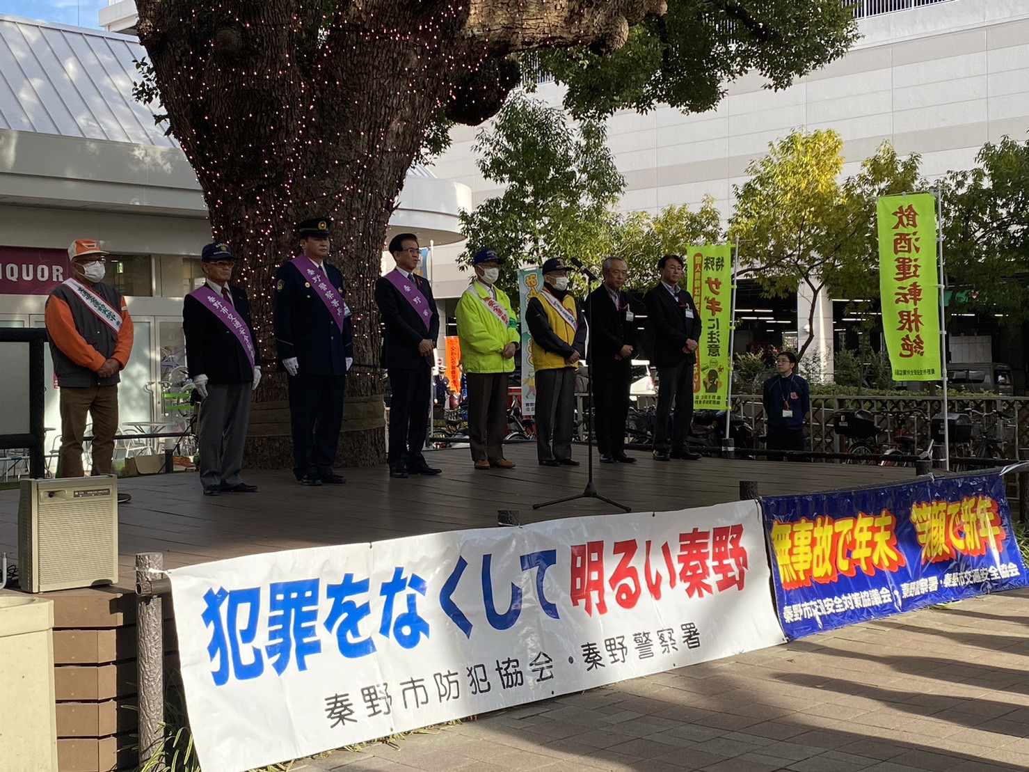 交通安全や防犯を呼びかける黄色いのぼりが立てられ、『犯罪をなくして明るい秦野』と書かれた横断幕が掲げられている街頭のステージ上で、警察官や関係団体のメンバー、スーツ姿の関係者たちが並んでいる交通事故防止・防犯キャンペーンの様子の写真
