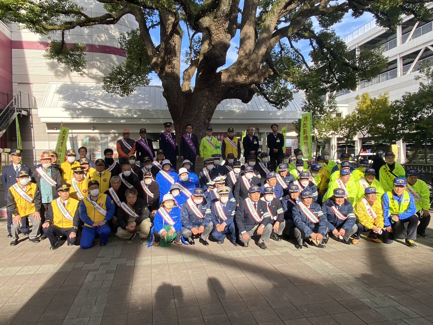 交通安全や防犯を呼びかける黄色いのぼりが立てられた屋外の広場で、大きな木の前に警察官、防犯パトロール隊、地域ボランティア、市職員など多くの関係者が制服やジャンパー、タスキを着用して整列・集合写真を撮っている交通事故防止・防犯キャンペーンの様子の写真