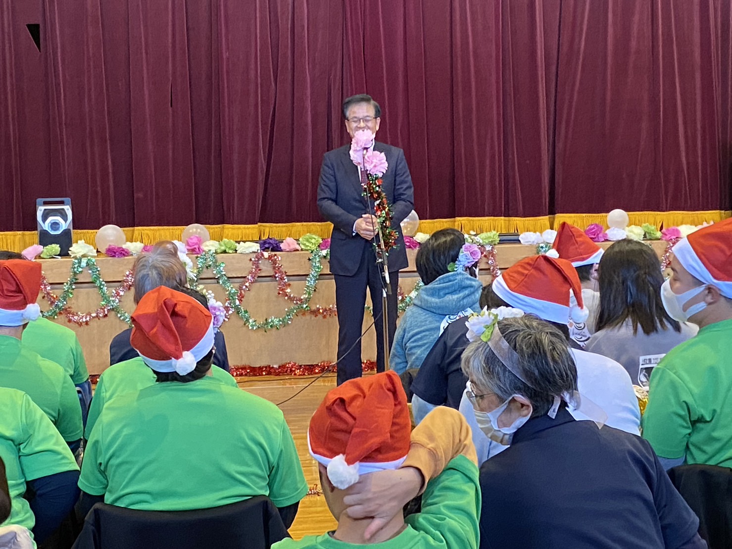 クリスマスの飾り付けがされた会場で、市長がマイクスタンドで話をしており、サンタ帽子をかぶり緑のTシャツを着て座っている参加者の方々が市長の話を聞いている写真