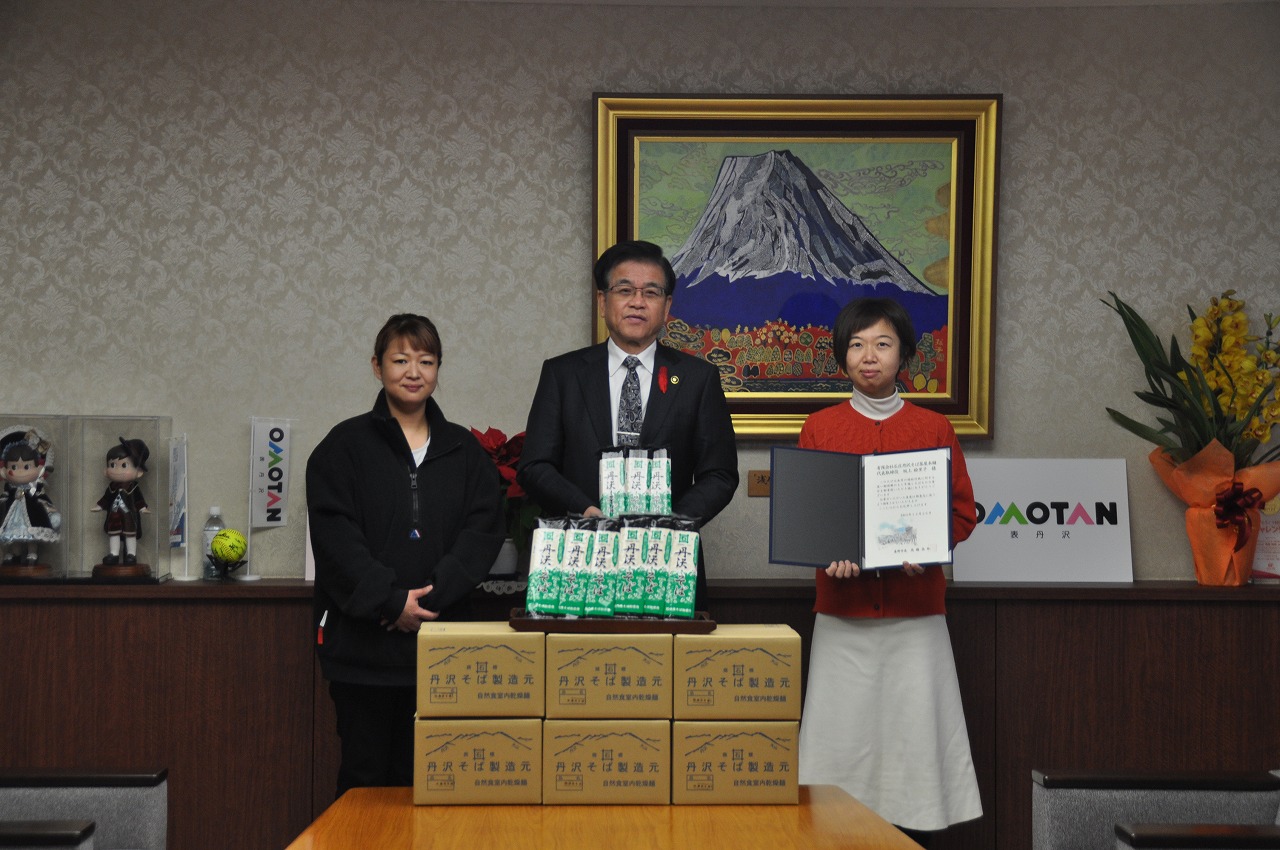 富士山の絵を背景に市長と関係者の二人の女性が並び、有限会社石庄丹沢そば茶屋本舗元から寄贈された多数のそば商品が並べられた机の前で感謝状を前にして記念撮影している様子の写真