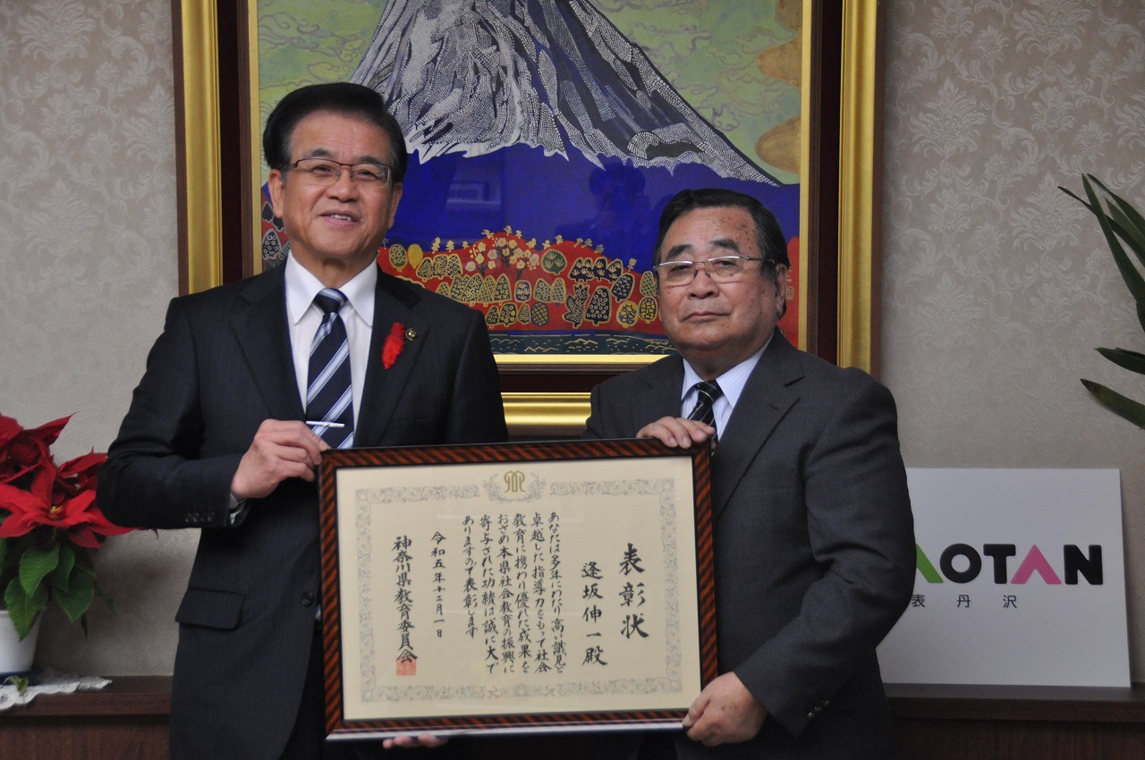 富士山の絵を背景に市長と逢坂伸一さんが、額に入った賞状をカメラに向けて掲げている受賞の記念写真
