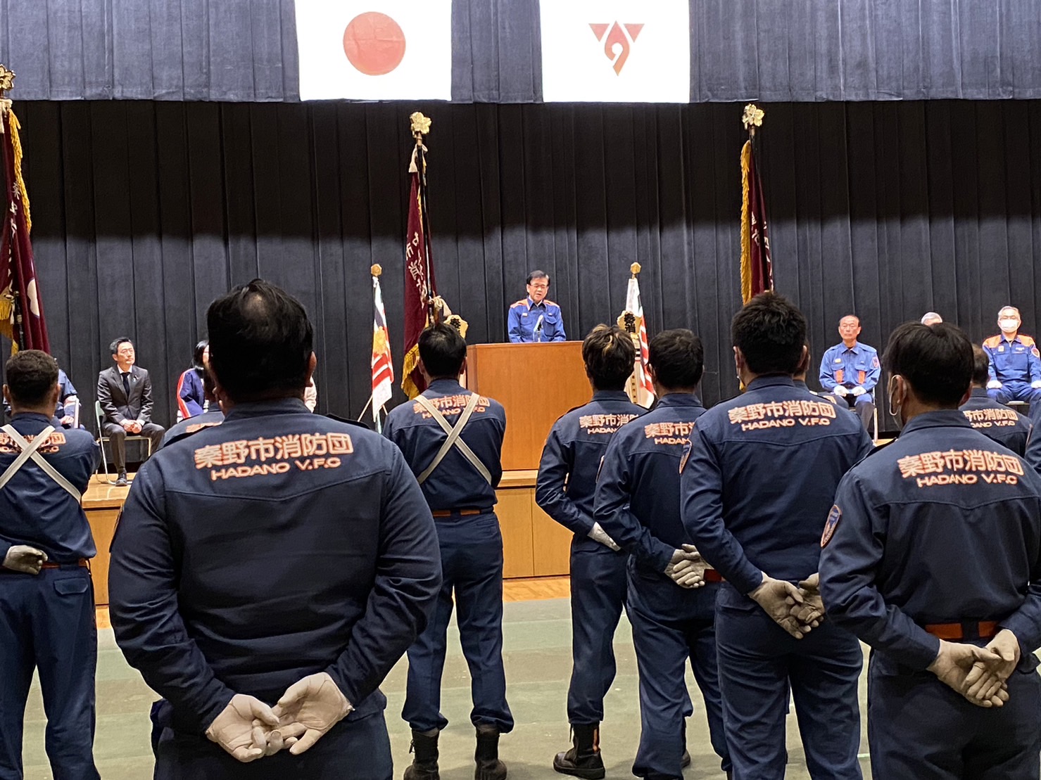 日本国旗と秦野市の市章が掲げられたステージに関係者が並び、その中央の演台で一人の関係者が話をしており、制服を着た団員たちが整列して話を聞いている激励式の様子の写真