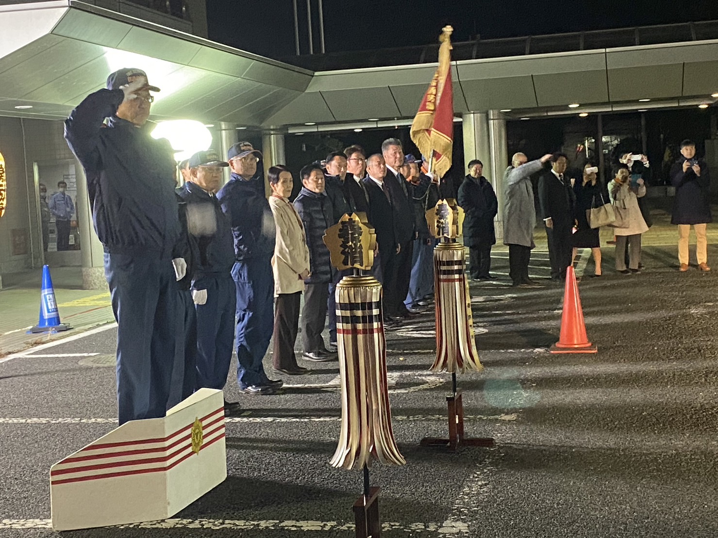 建物の前で整列した制服姿の消防団員と関係者たちが敬礼し、纏（まとい）や団旗が掲げられ、周囲で市民や関係者が見守る夜間の激励式の様子の写真