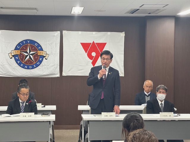 「CITY OF PASADENA」と「秦野市旗」の旗が掲げられた会場で、高橋市長がマイクを持って話をしている様子の写真