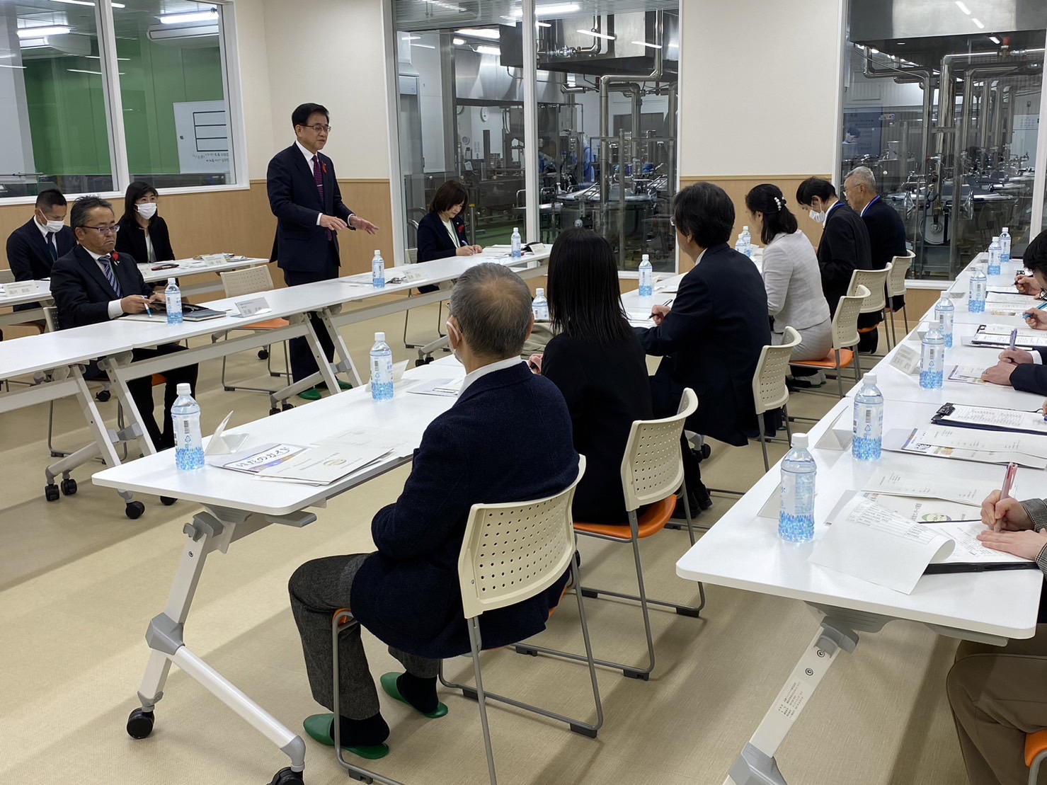 秦野市の関係者と座間市の関係者の方々が向かい合う形で席に着き、高橋市長が立って話をしており、関係者の方々が座って資料に目を通しながら話を聞いている様子の写真