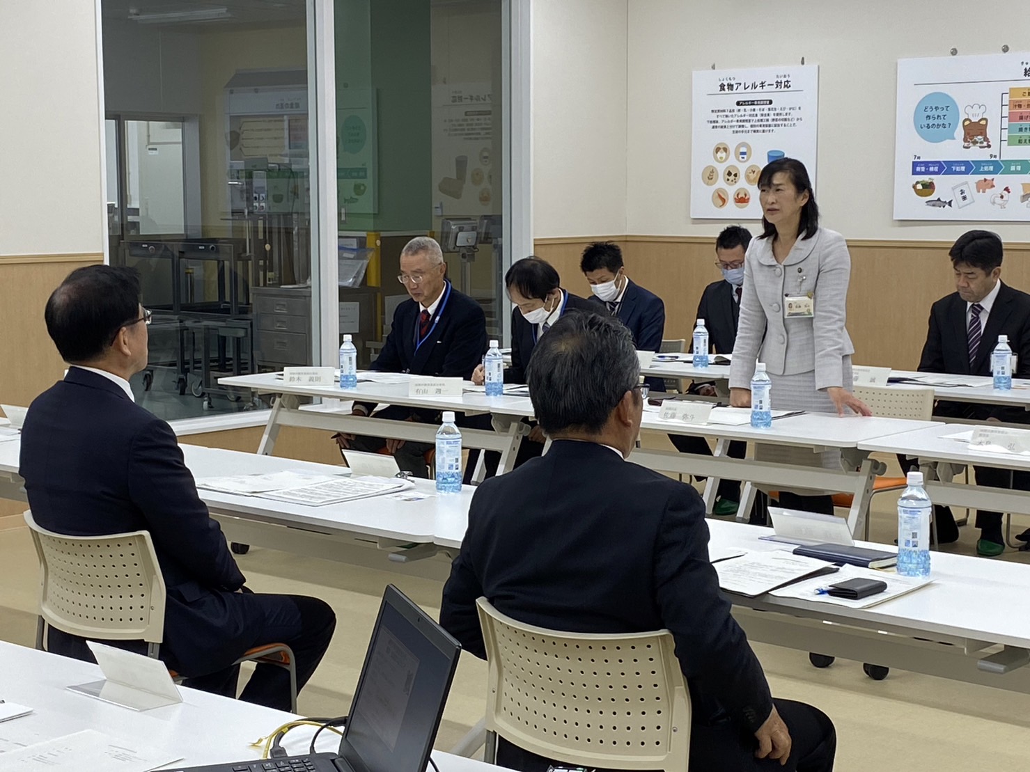 秦野市の関係者と座間市の関係者の方々が向かい合う形で席に着き、女性が立って話をしており、関係者の方々が座って資料に目を通しながら話を聞いている様子の写真