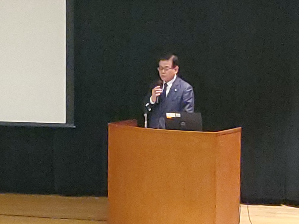 檀上にある演題の前にに立ち、マイクを持って話すスーツ姿の男性を写した写真