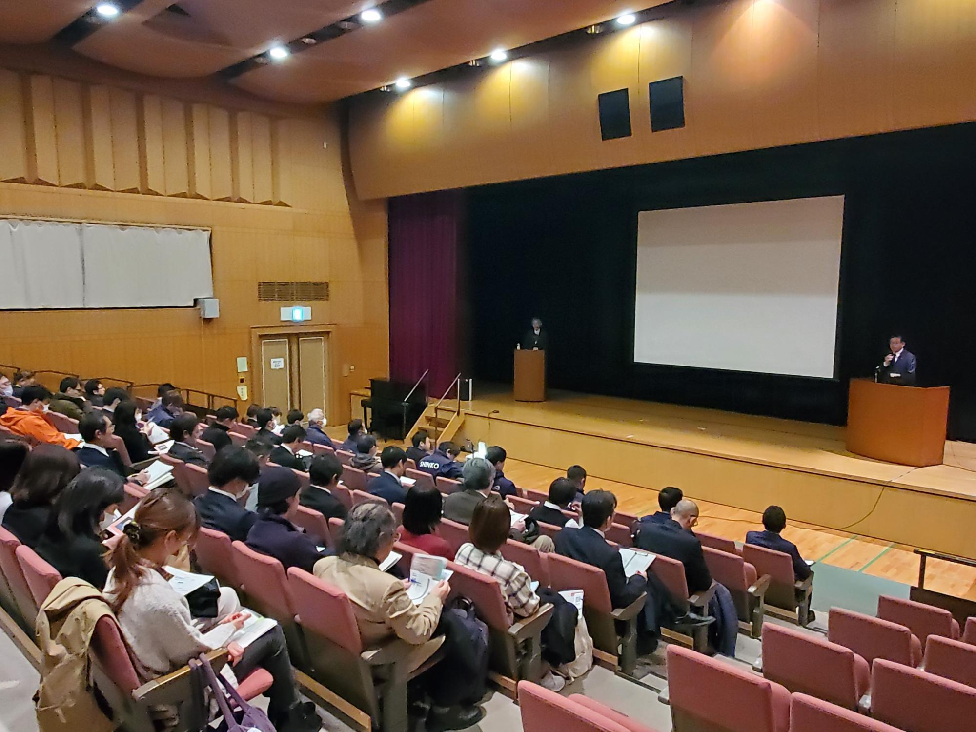 会場の舞台には大きなスクリーンが設置され、演台に立つ講師の男性が話しており、客席には多くの参加者が座り配布資料を見ながら話を聞いている様子が写っている画像の写真