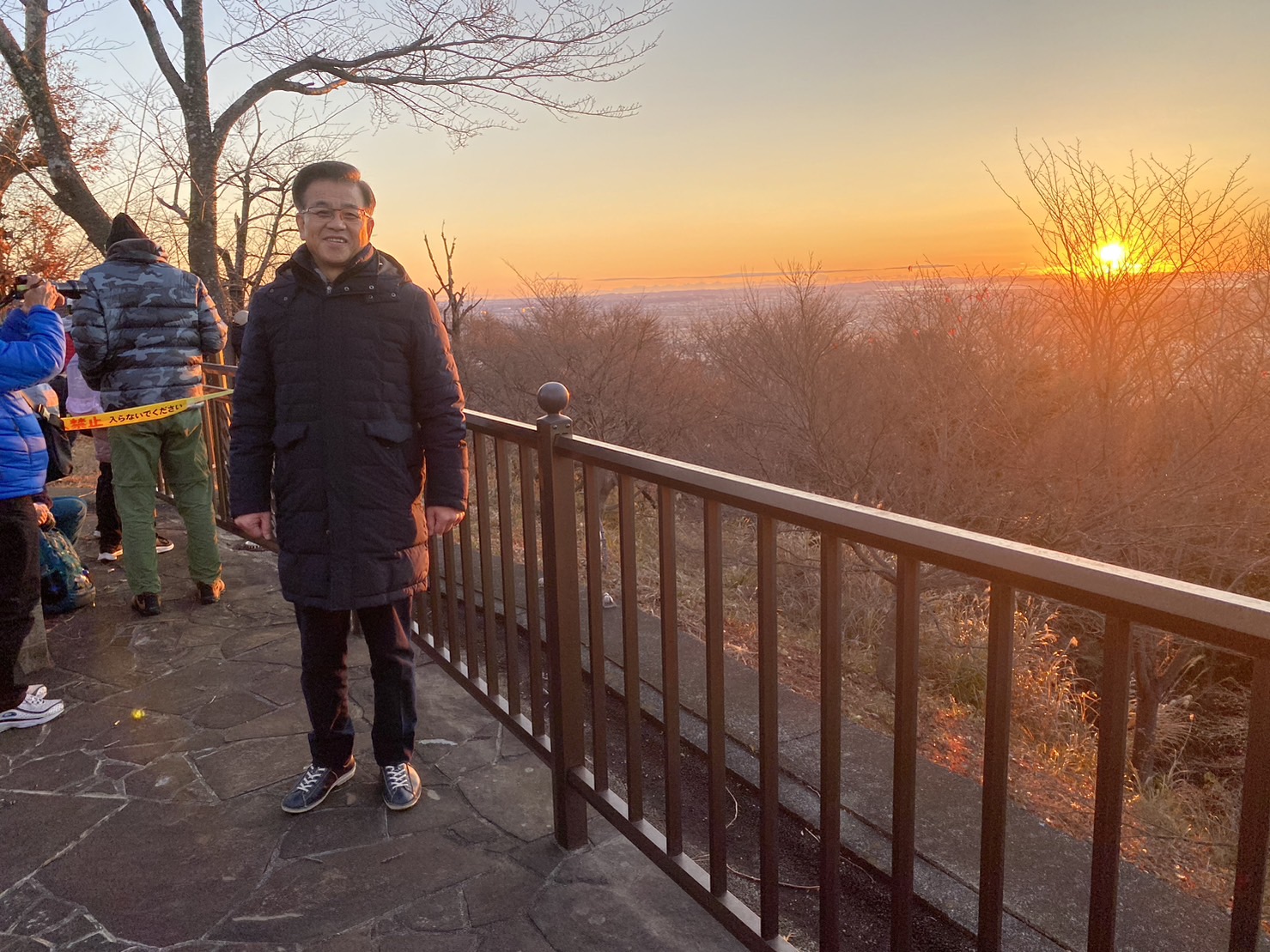朝日が昇る山の風景を背に黒いコート姿の市長が展望台で立ち、周囲に初日の出を見守る人々がいる写真