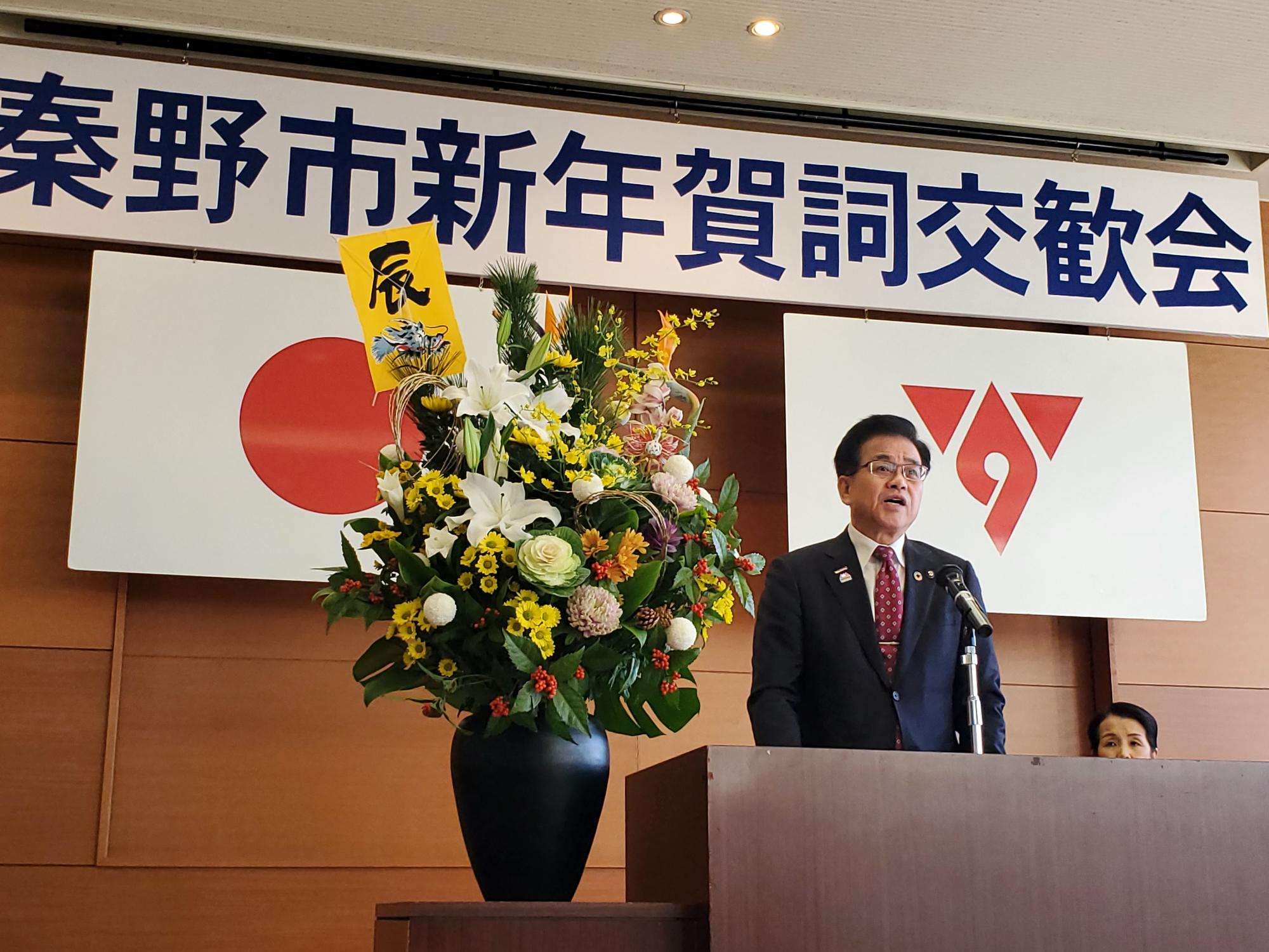 日の丸と市の旗が掲げられている壇上でスーツ姿の市長がマイクを手に新年賀詞交歓会の挨拶を行っている写真