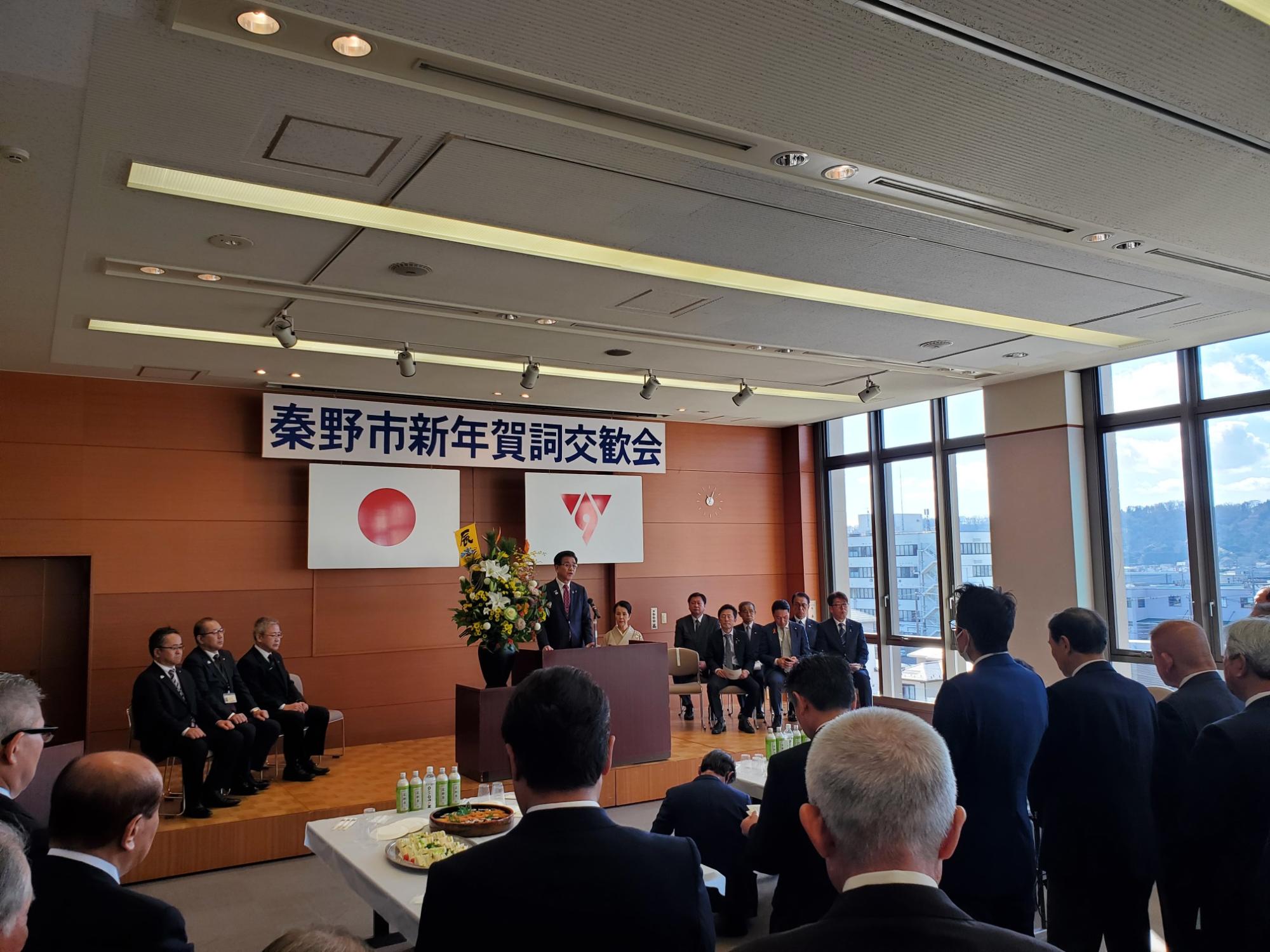 秦野市新年賀詞交歓会の会場で壇上の市長が挨拶し、周囲に来賓や出席者が並び、手前に料理が並ぶ様子を後方から写した写真