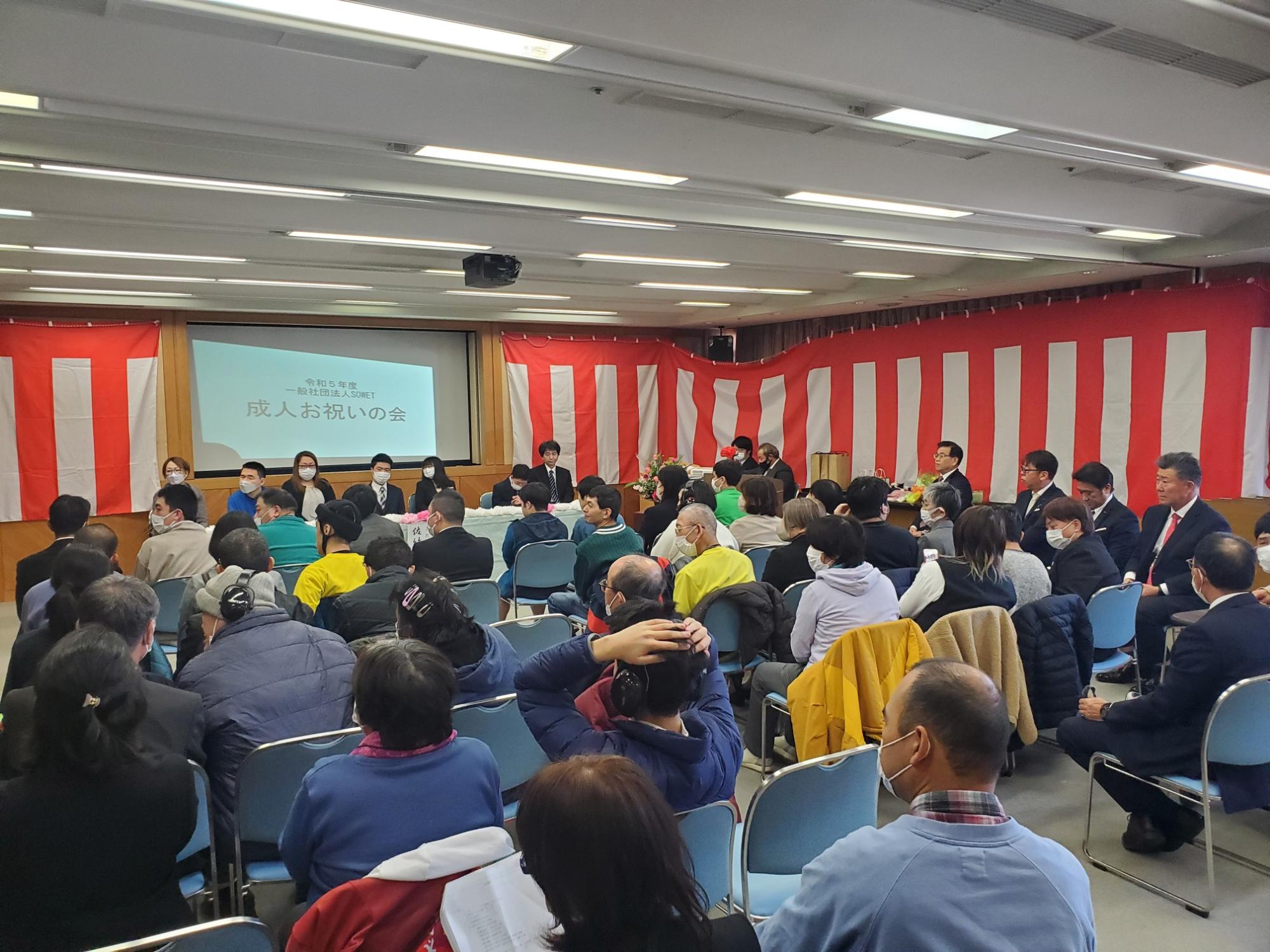 会場に多くの人が集まり前方のスクリーンに「成人お祝いの会」と映し出されている成人お祝いの会の会場の写真