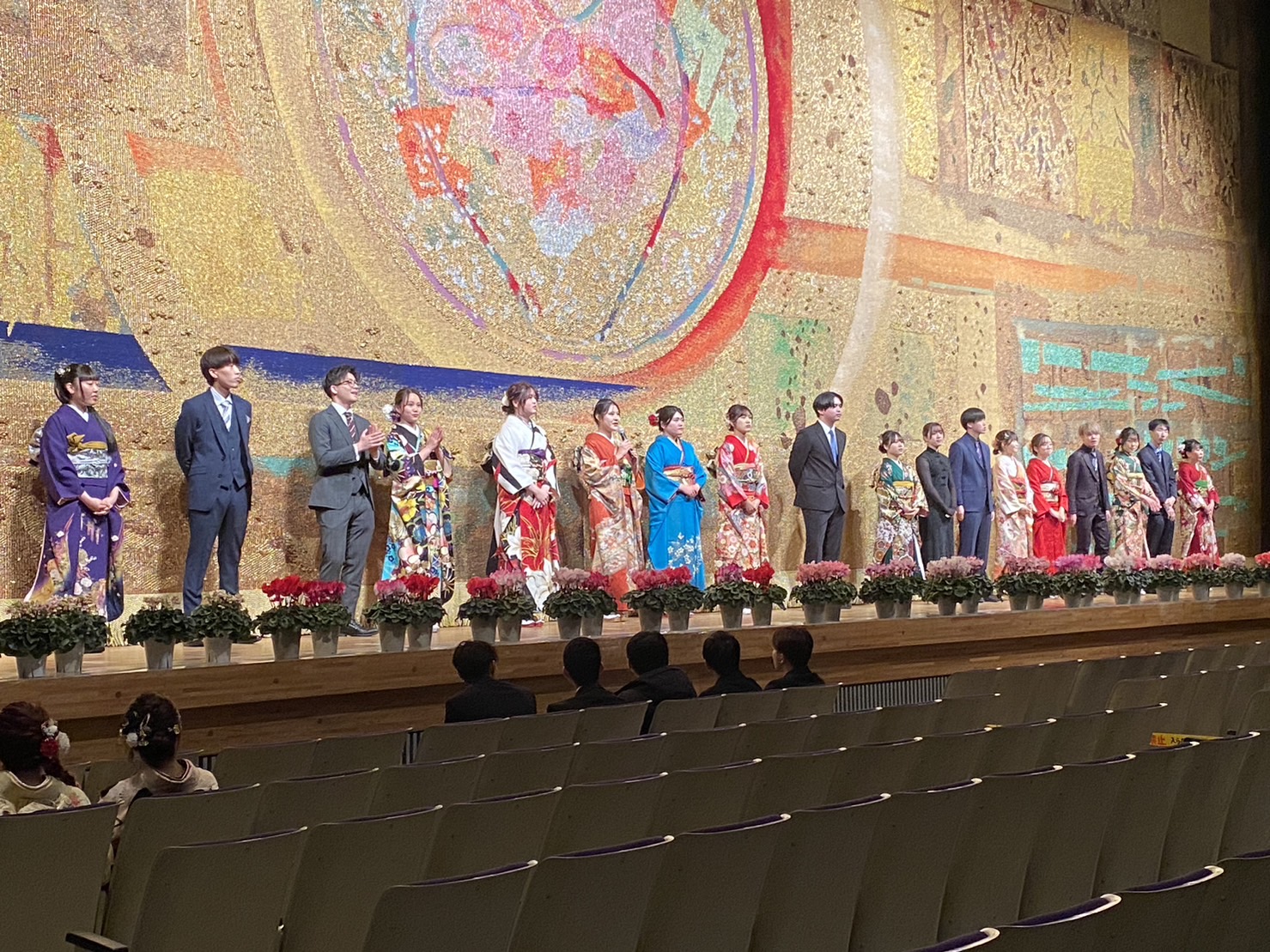 金色の豪華な幕の前で、華やかな振袖やスーツ姿の若者たちが横一列に整列し、振袖姿の女性がマイクを持ち客席に向かい話をしている様子の写真