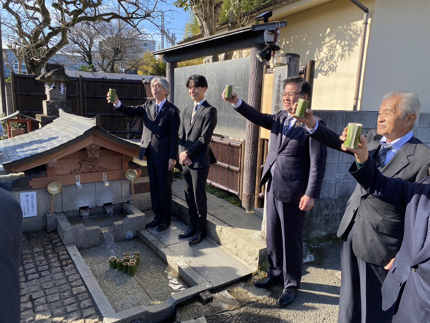 屋外の湧水施設前でスーツ姿の男性4人が竹筒の杯を掲げて乾杯のように並んでいる写真