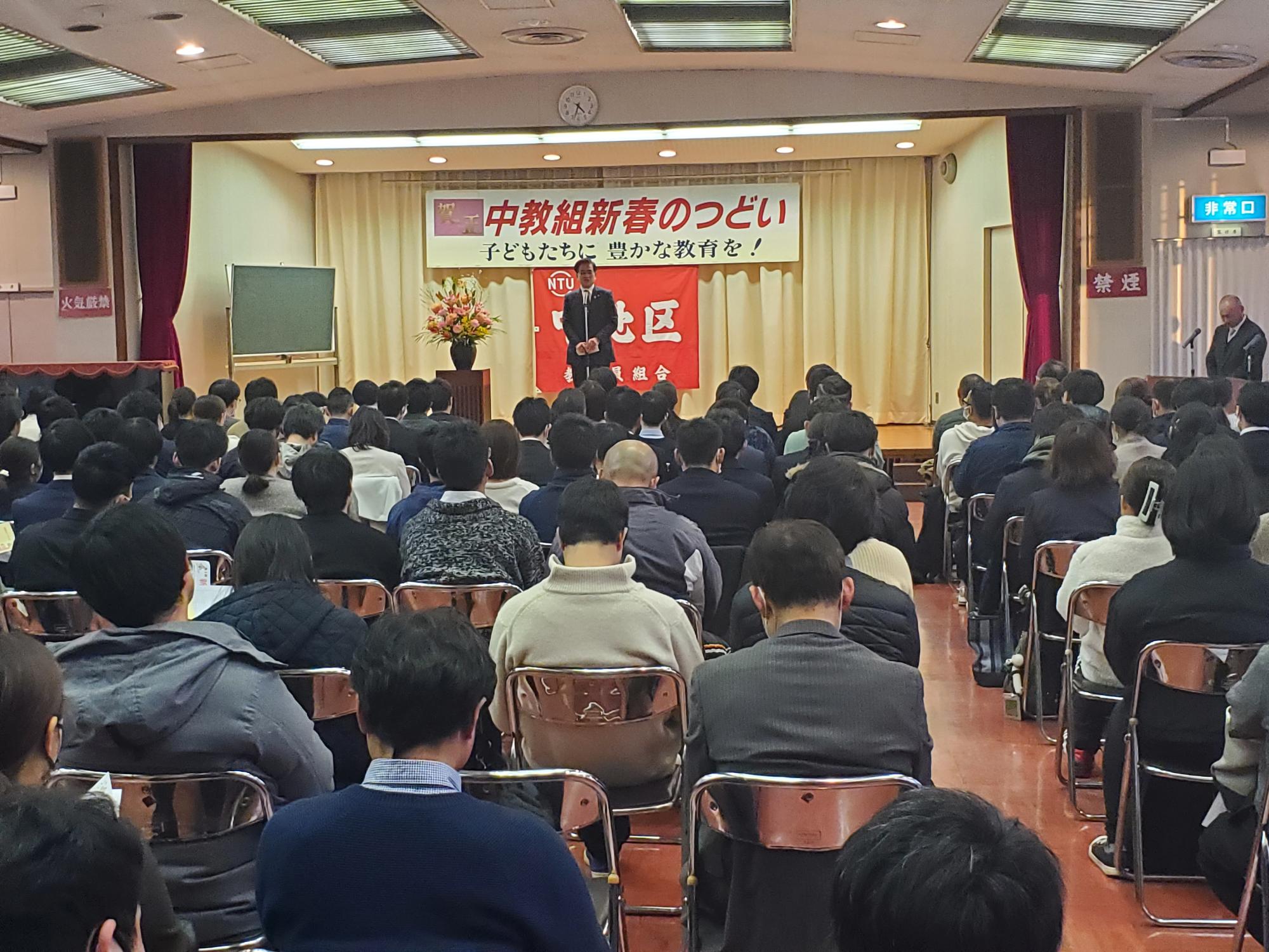 「中教組新春のつどい」と書かれた横断幕の前に立ち話をする市長と会場に多くの参加者が集まった中地区教職員組合新春のつどいの写真