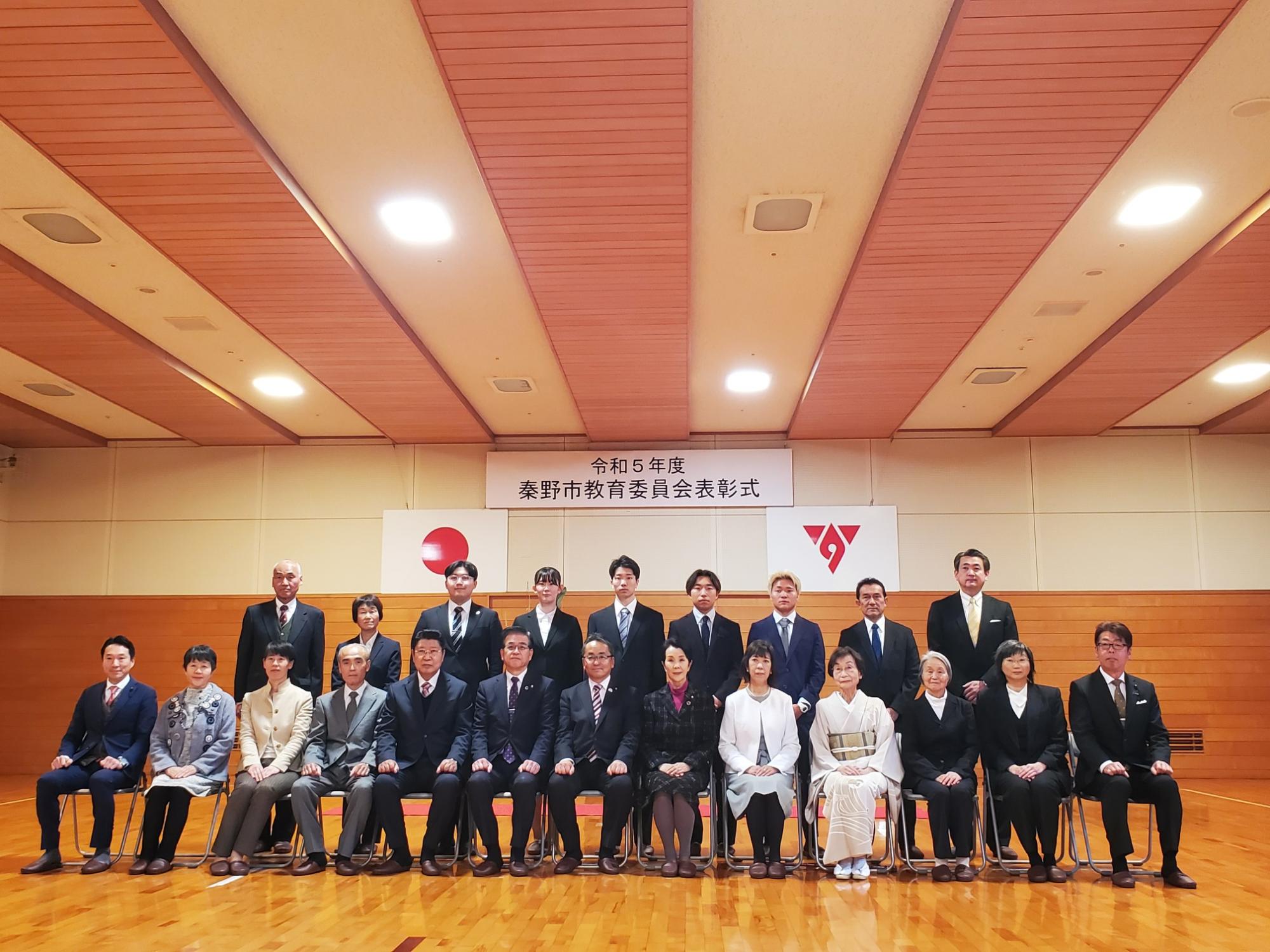 体育館で行われた令和5年度秦野市教育委員会表彰式で、受賞者と関係者が横一列に並び記念撮影する写真