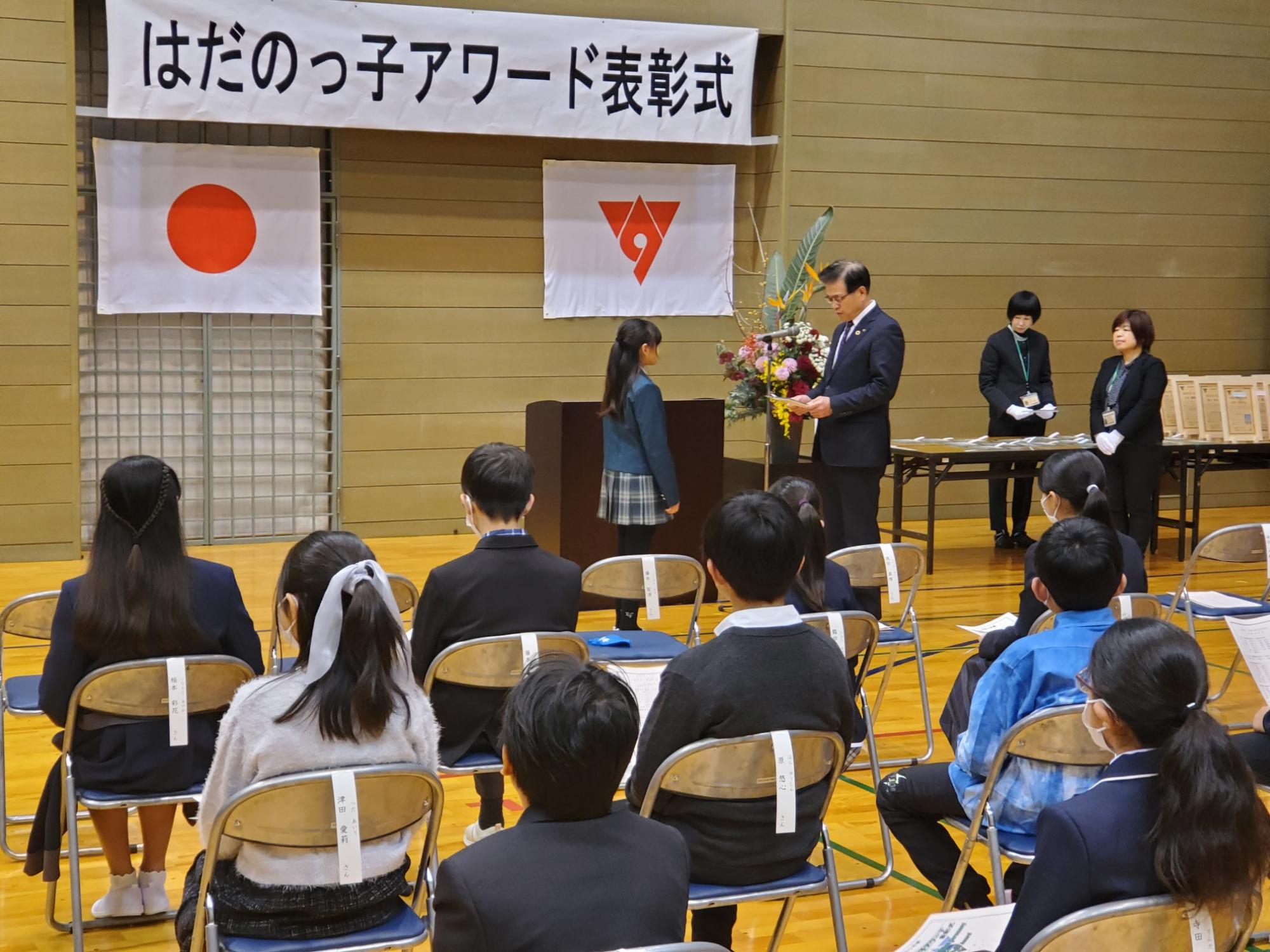 体育館で行われた「はだのっ子アワード表彰式」で、壇上の前で市長が女子児童に賞状を渡している様子の写真