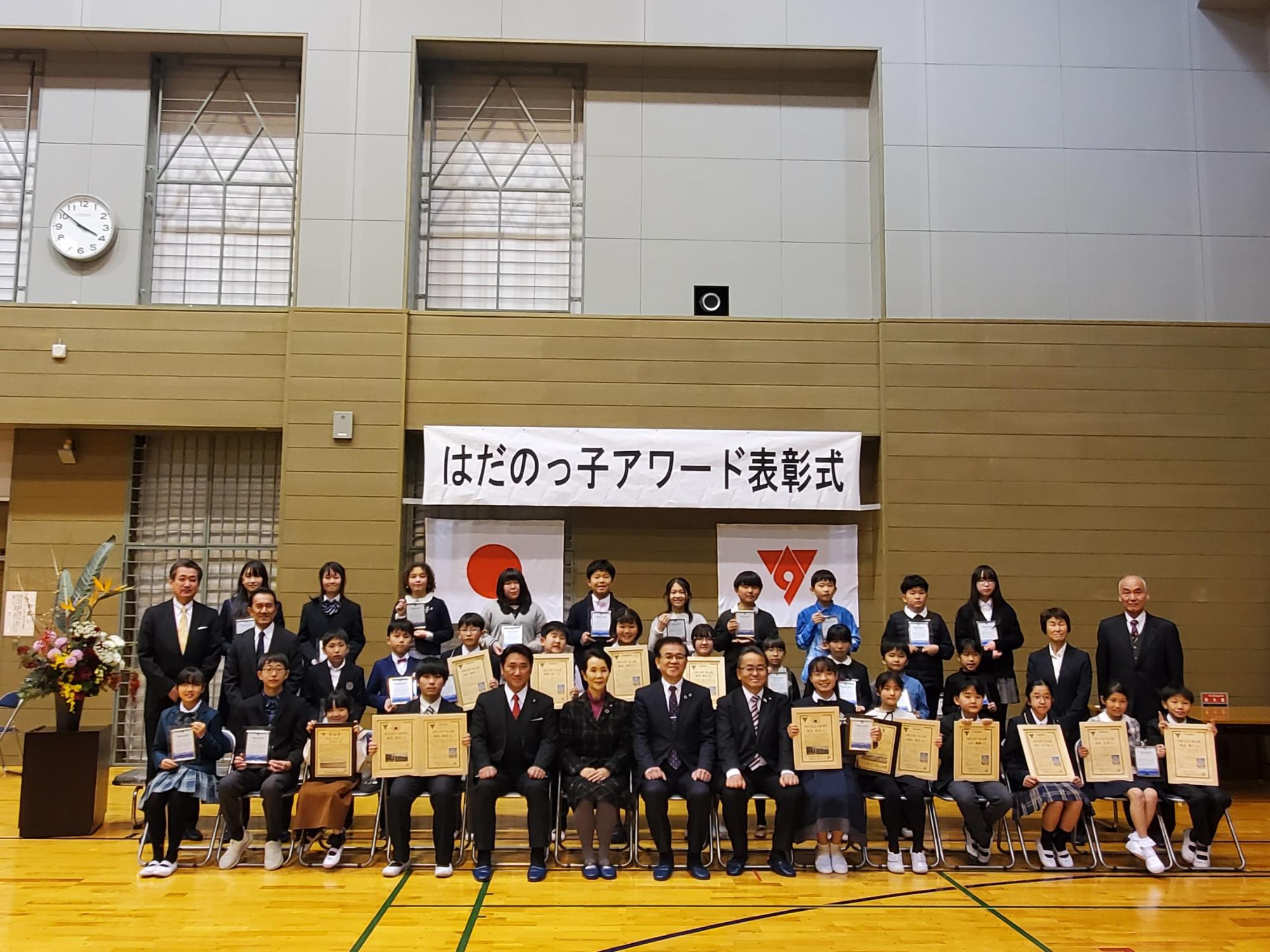 体育館で行われた「はだのっ子アワード表彰式」で、賞状や盾を手にした子どもたちと関係者が横一列に並び記念撮影する写真
