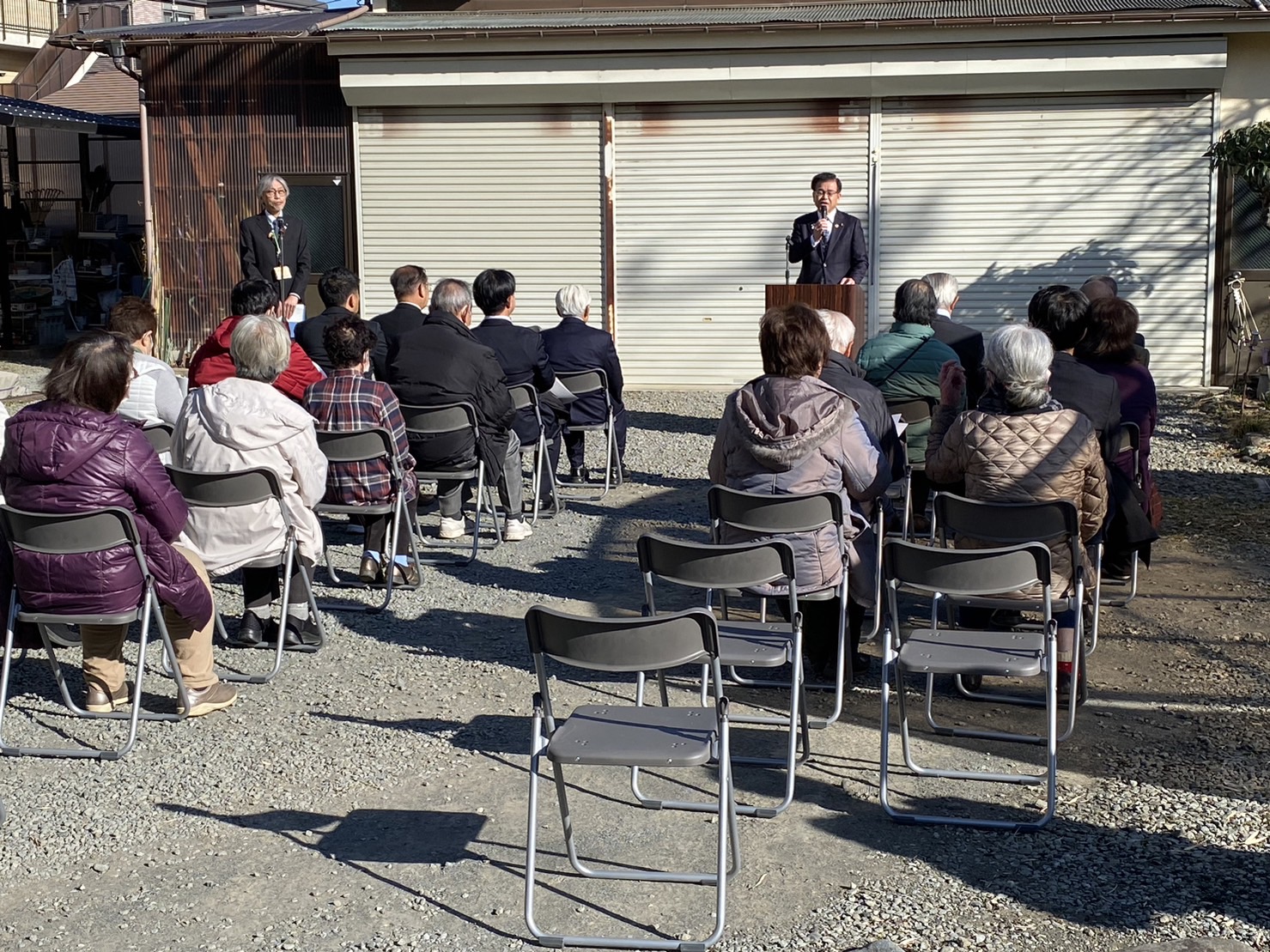 屋外の簡易な会場で折りたたみ椅子に座る人々を前に、スーツ姿の市長が演台でマイクを持ち挨拶している写真