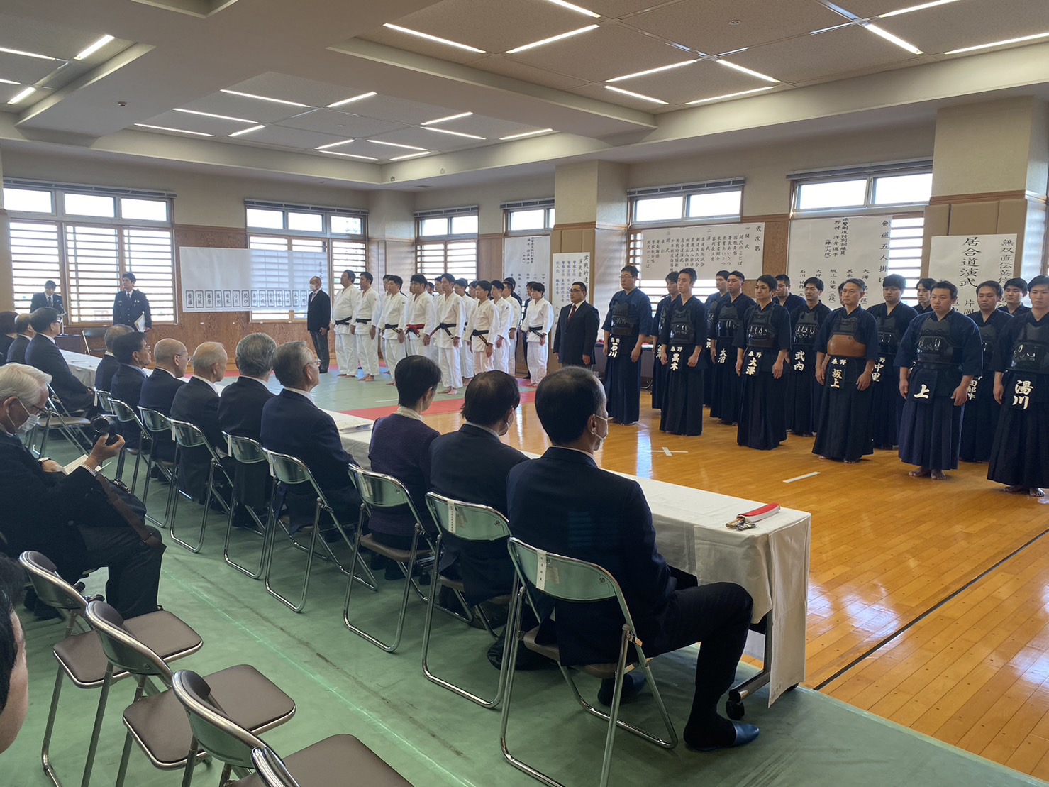 体育館の緑色のシートの上で柔道着姿と剣道防具姿の選手が横一列に並び、前列にはスーツ姿の来賓が座る式典の様子を写した写真
