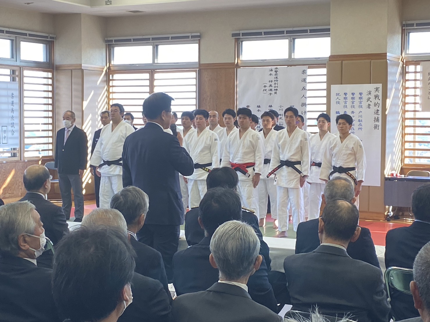 柔道着を着た選手たちが整列し、前方ではスーツ姿の市長がマイクを持って話し、観客席には来賓が並ぶ式典風景の写真