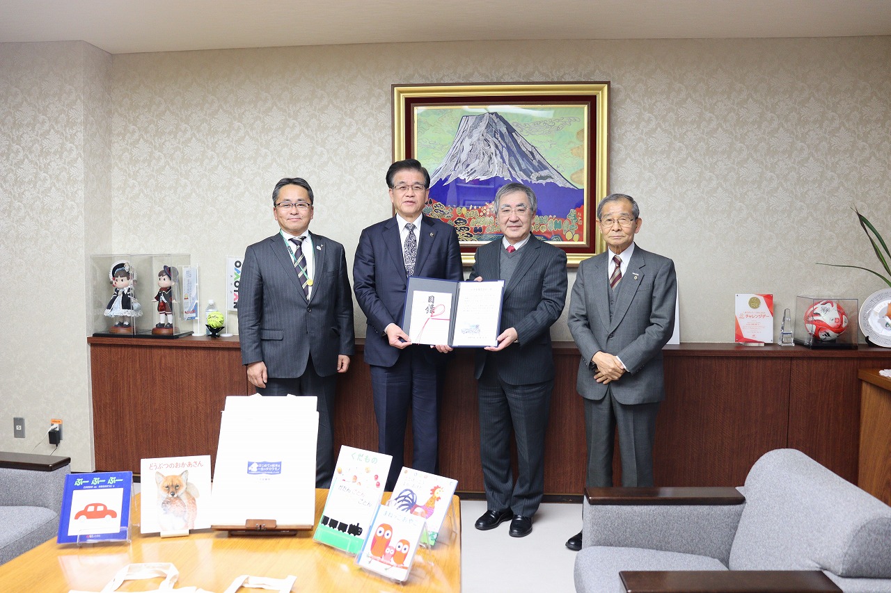 十全堂株式会社の方々と市長が横一列に並び、市長と中央の男性が賞状を持ち、背後には富士山の絵画や飾り物、手前の机には絵本が置かれた写真