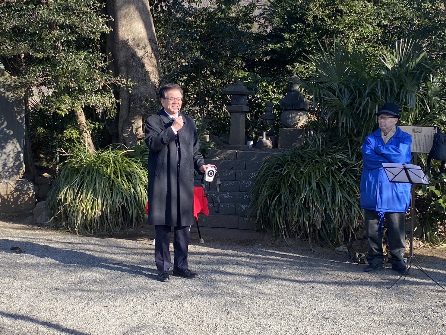 屋外の神社境内で黒いコート姿の市長がマイクを手に話し、青い上着の男性が隣に立ち話を聞いている写真