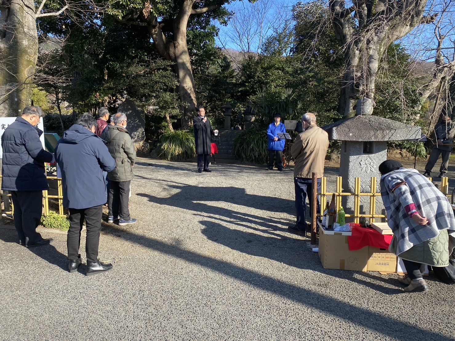 神社境内で黒いコート姿の市長がマイクで話し、青い上着の男性や参列者数名が立ち話を聞いている様子を写した写真
