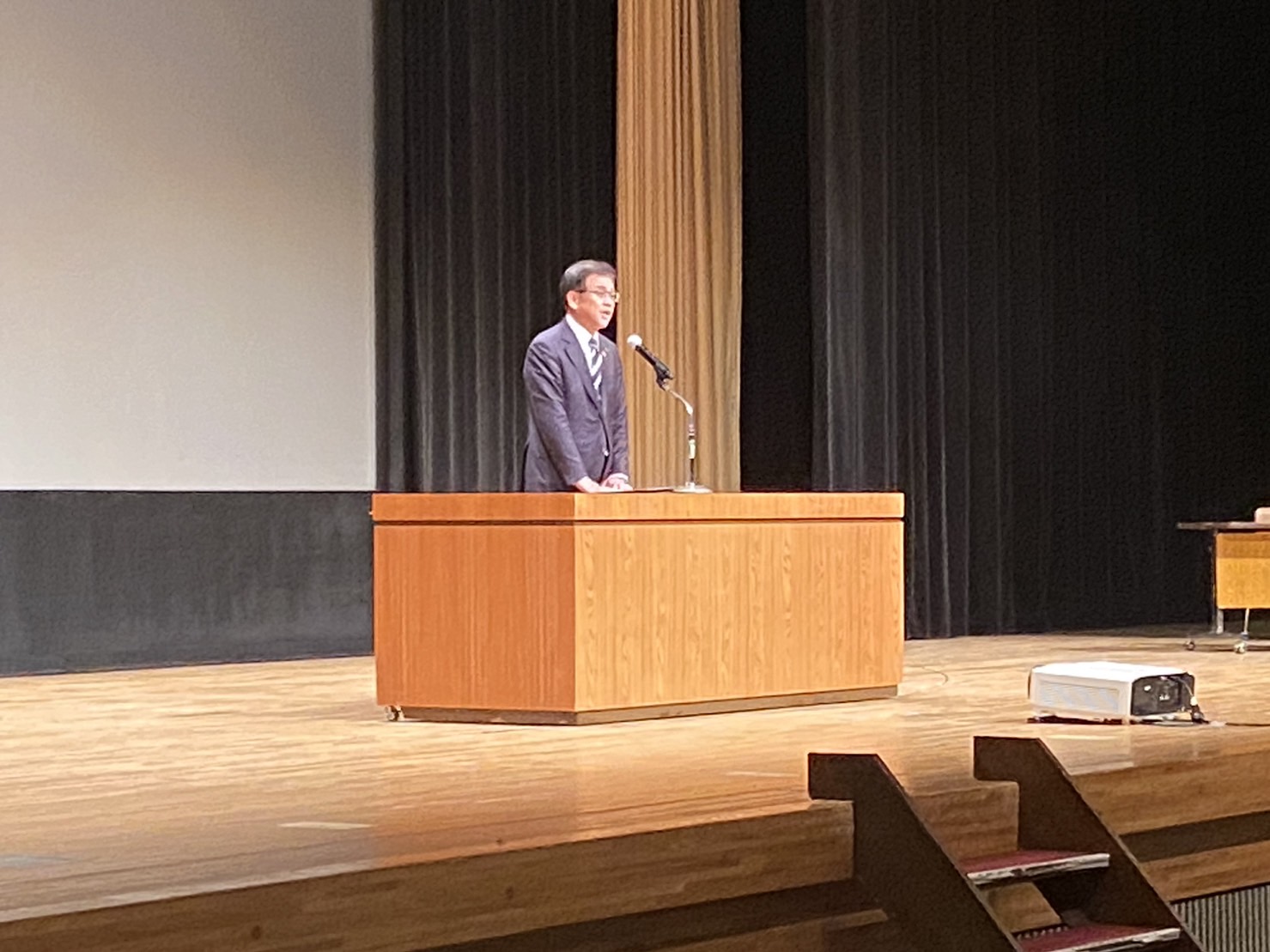 大きな舞台上でスーツ姿の市長が演台に立ちマイクを使って挨拶する様子を写した写真