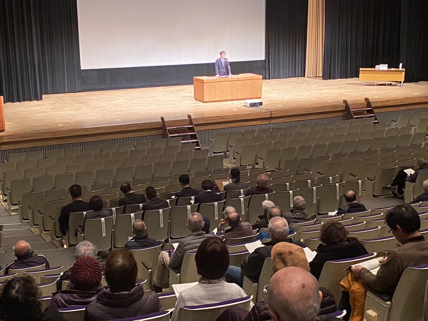 広い講堂でスーツ姿の市長が演台から話し観客席には関係者や参加者の方々がを着席している様子を左後方から写した写真