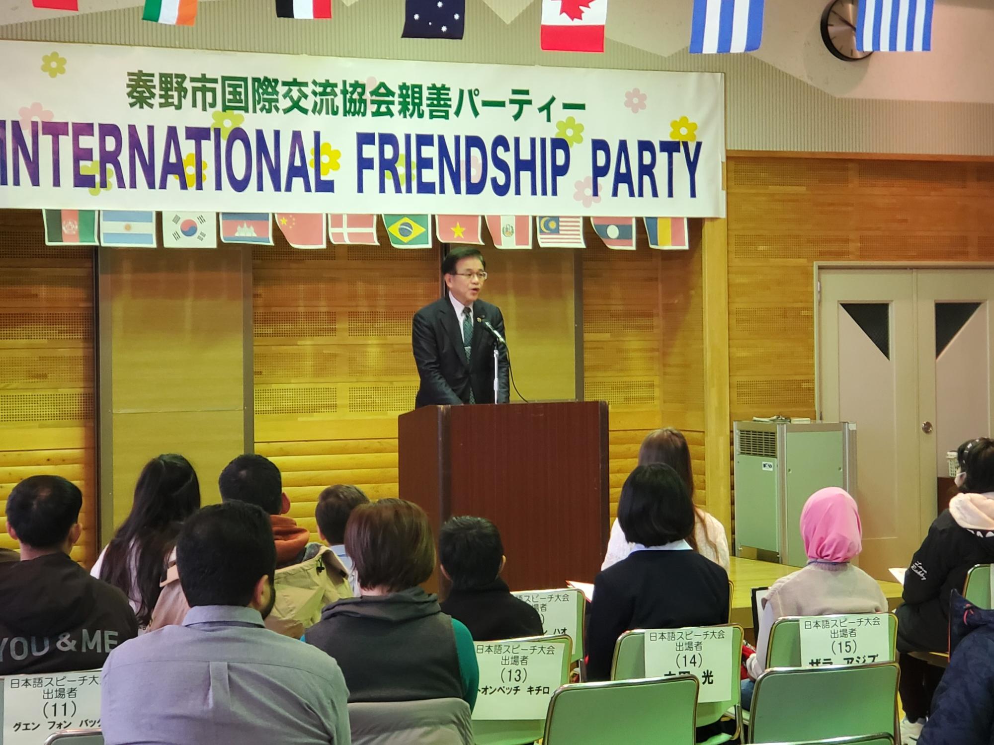 国際交流イベント会場で演台に立つ市長がスーツ姿で挨拶し、観客が椅子に座って話を聞いている様子の写真