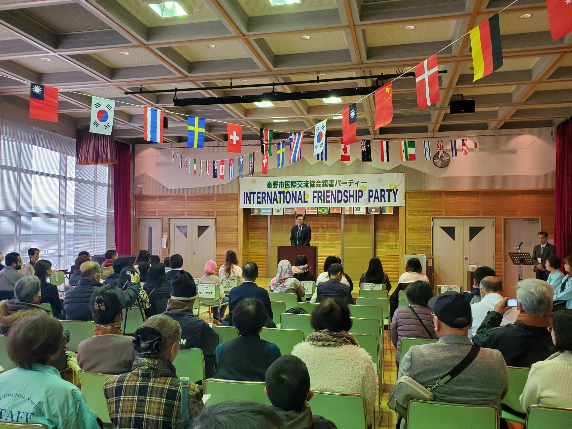 国際交流パーティー会場で演台に立つ男性が挨拶し、客席には多くの参加者が座り天井には各国の国旗が並ぶ国際交流フェスティバル全体の写真