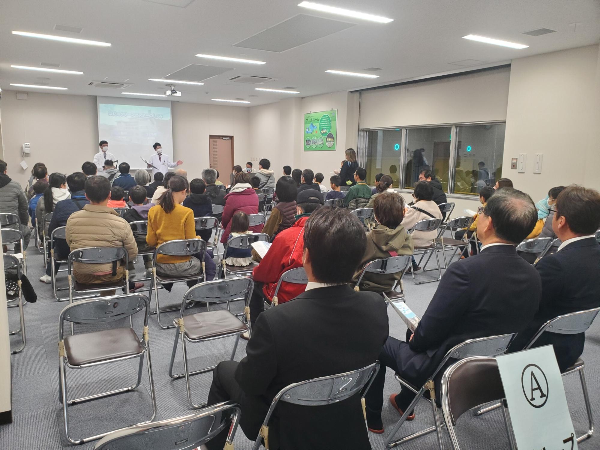 多数の参加者が椅子に座り、前方で白衣を着た二人がプレゼンを行う様子を写した室内イベントの写真