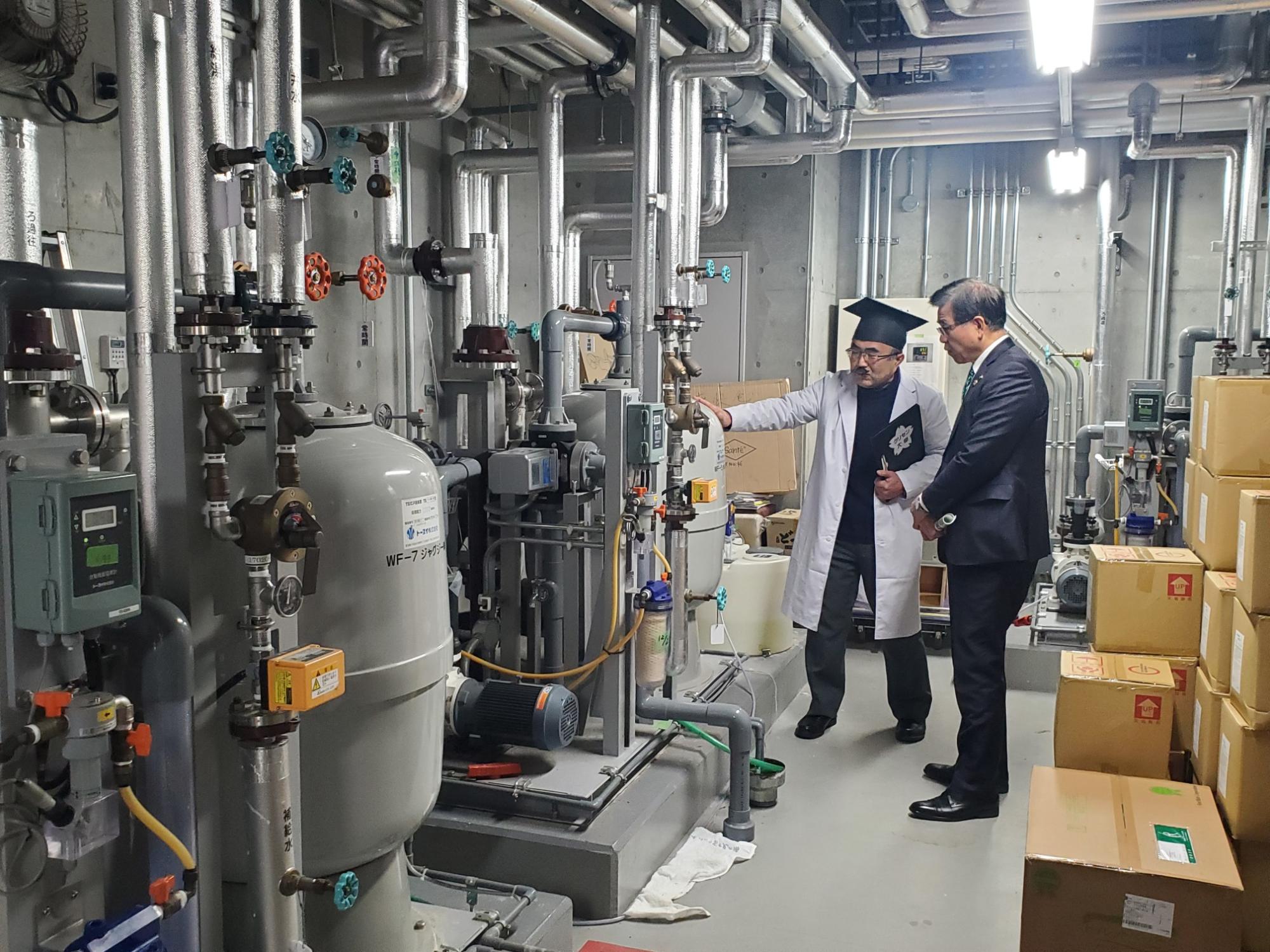 配管やタンクが並ぶ機械室で白衣の男性がスーツ姿の市長に設備を指差し説明している様子の写真