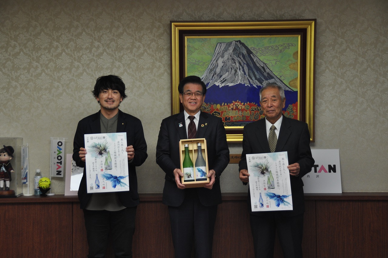 金井酒造店と株式会社大地が市長と一緒に並び、市長が日本酒の箱に入った瓶セットを掲げ、両脇の人物が同じデザインのポスターを持っている記念写真
