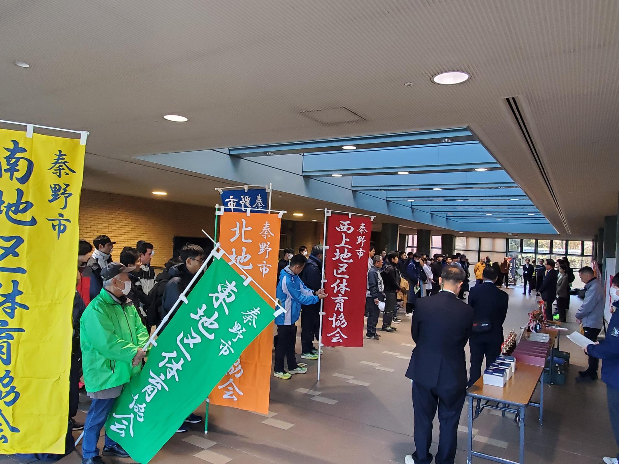 屋内会場で秦野市の各地区体育協会が色鮮やかな旗を掲げて並び、前方に表彰用のトロフィーや賞品が並ぶ写真
