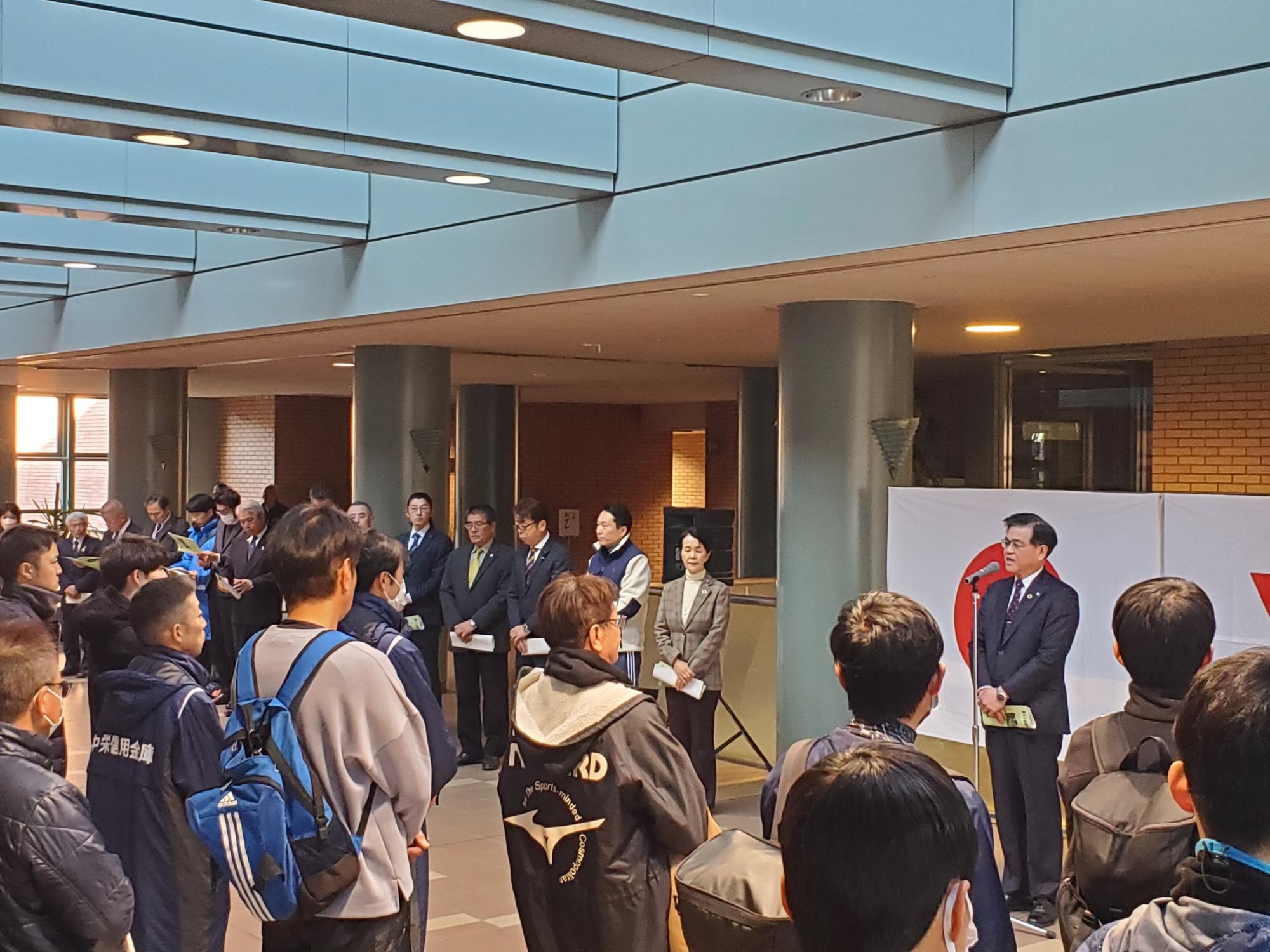 屋内会場でスーツ姿の市長が壇上でマイクを持ち挨拶し、背後に関係者が並び、前方に多くの参加者の方々が話を聞いている様子の写真