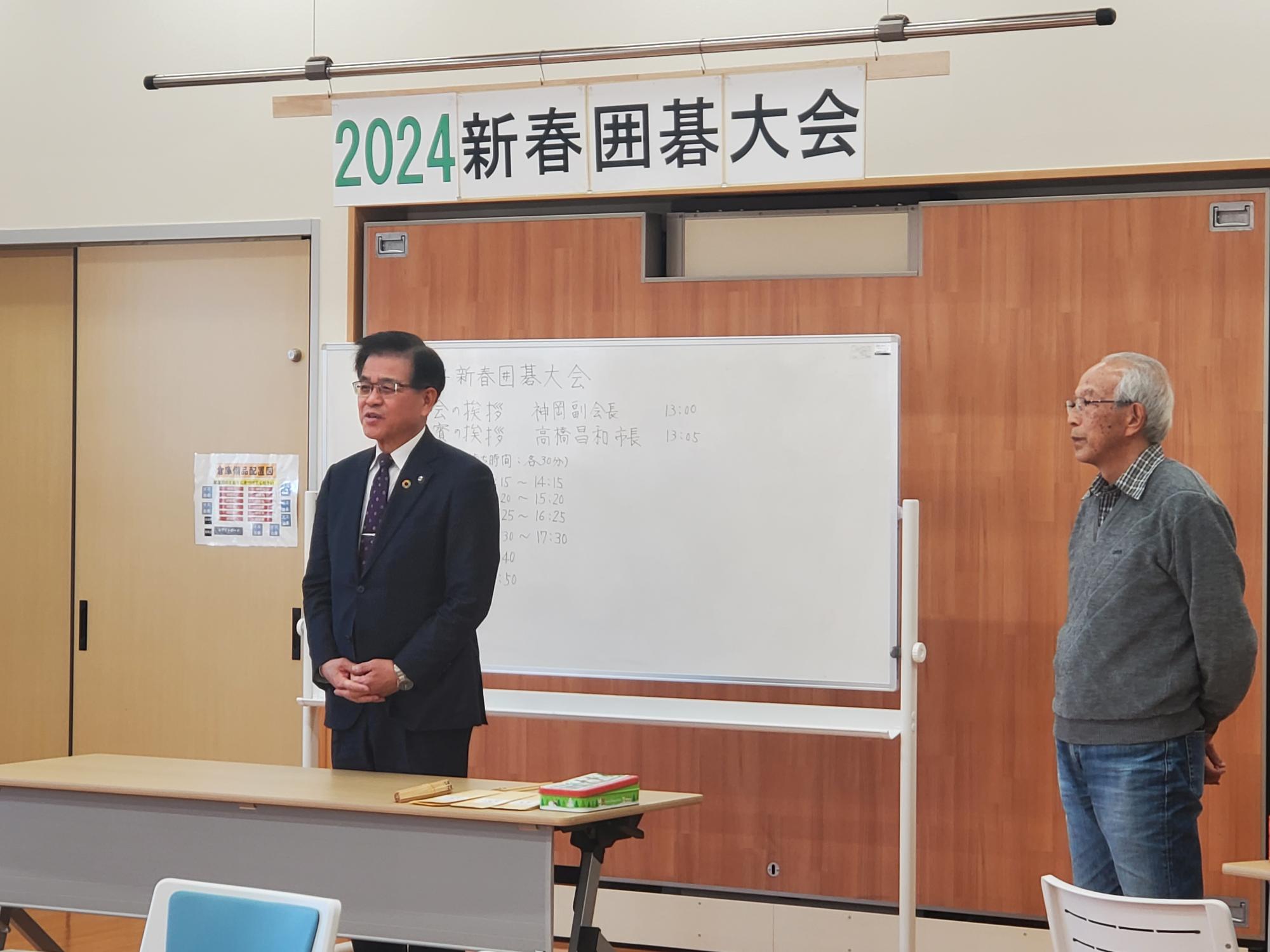 2024新春囲碁大会の会場でスーツ姿の市長が挨拶し、白板には進行予定が書かれ隣に年配男性が並ぶ写真