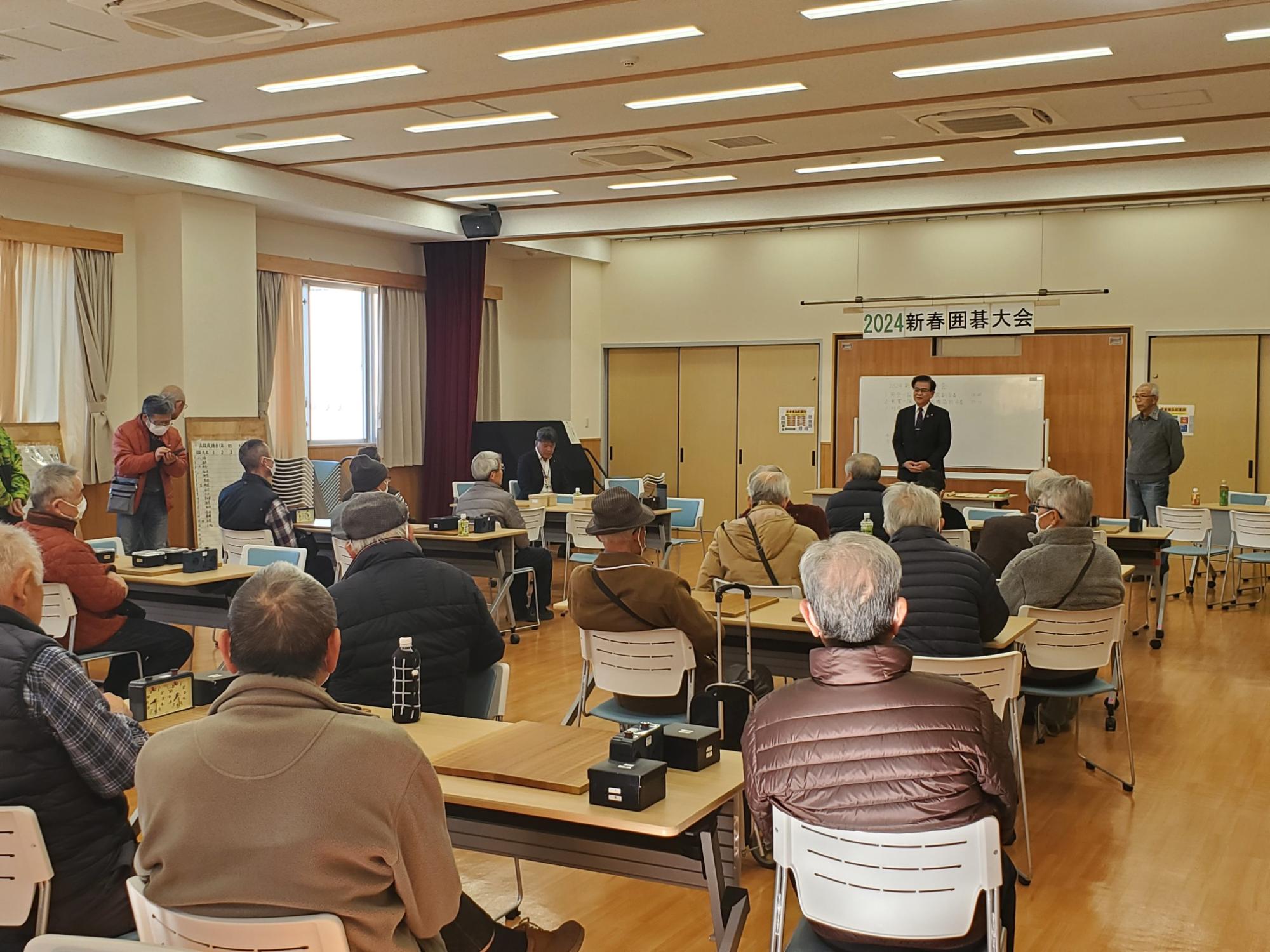 2024新春囲碁大会の会場で多くの参加者が椅子に座り、前方でスーツ姿の市長が挨拶している様子を後方から写した写真