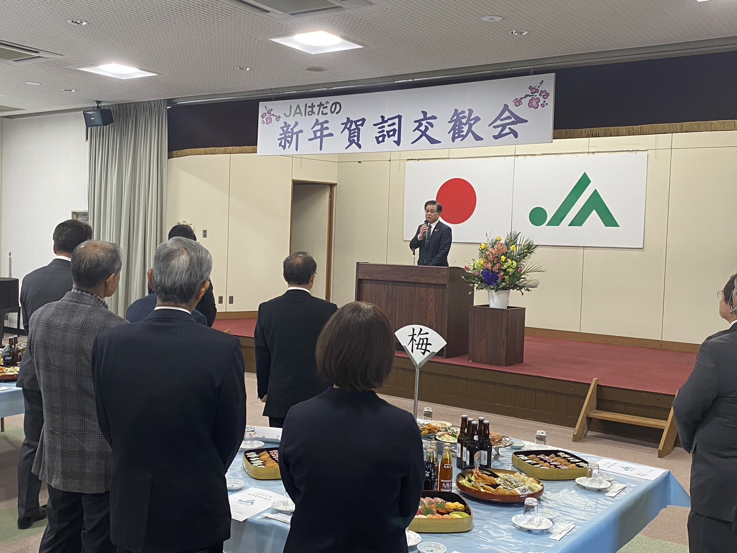 「JAはだの新年賀詞交歓会」で演台の市長が挨拶し、会場では立食形式の料理が並び参加者が起立して聞いている様子の写真