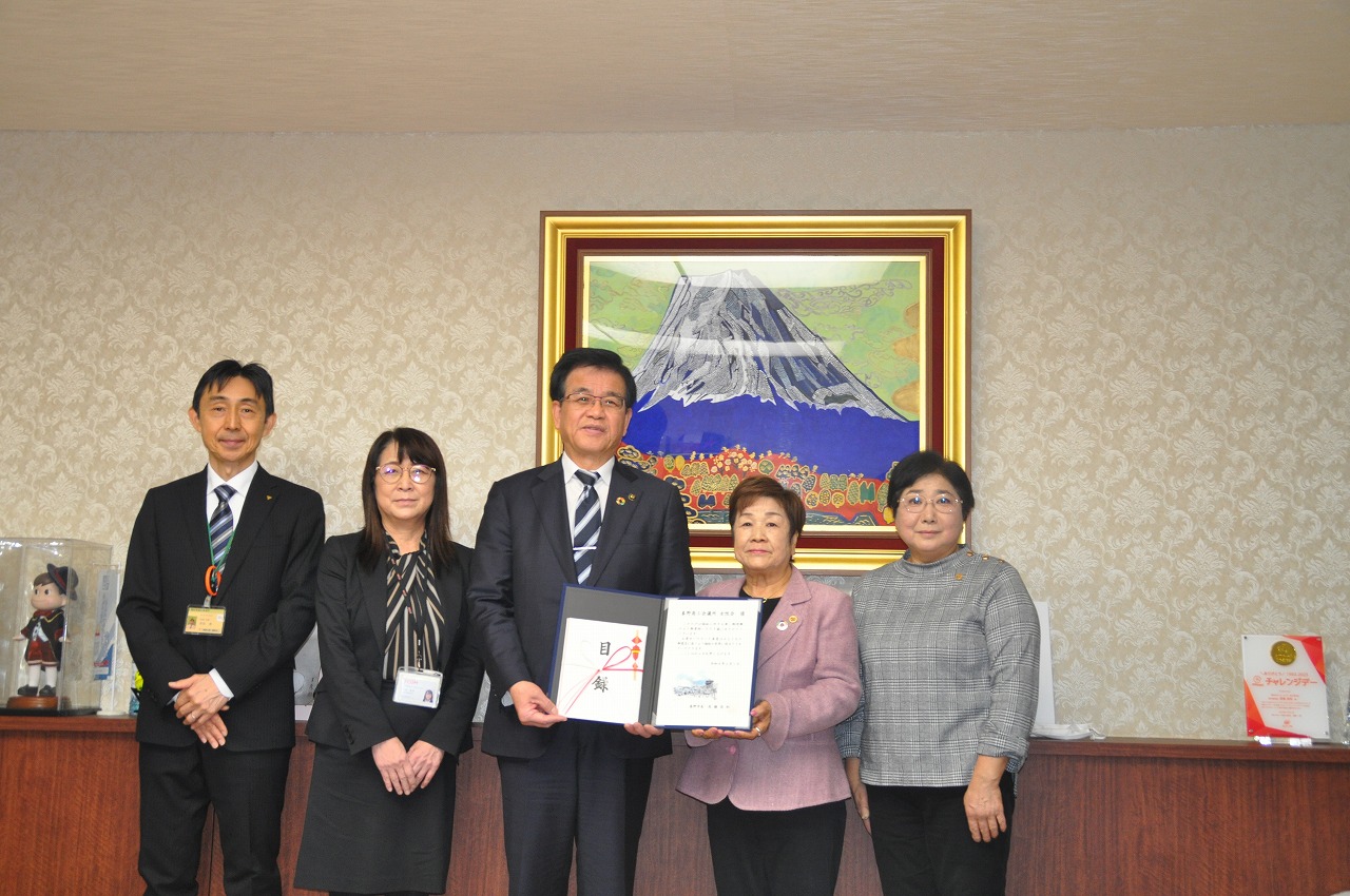 スーツ姿の市長と秦野商工会議所女性会の関係者の方々が横一列に並び、市長と中央の女性が賞状と贈呈品を掲げている記念写真