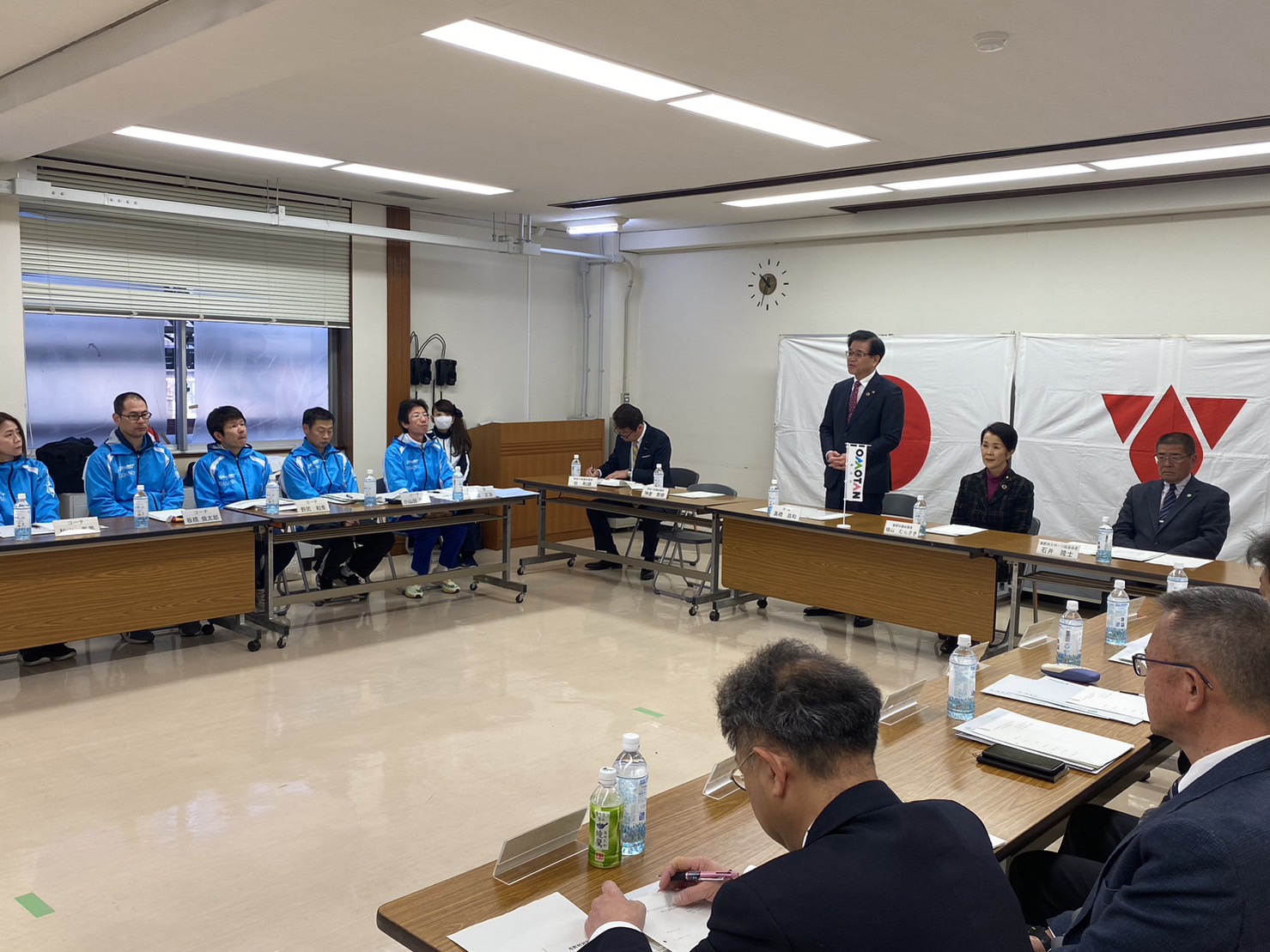 会議室でスーツ姿や青いジャンパー姿の人々が机を囲み、市長が立って話をしている様子の写真